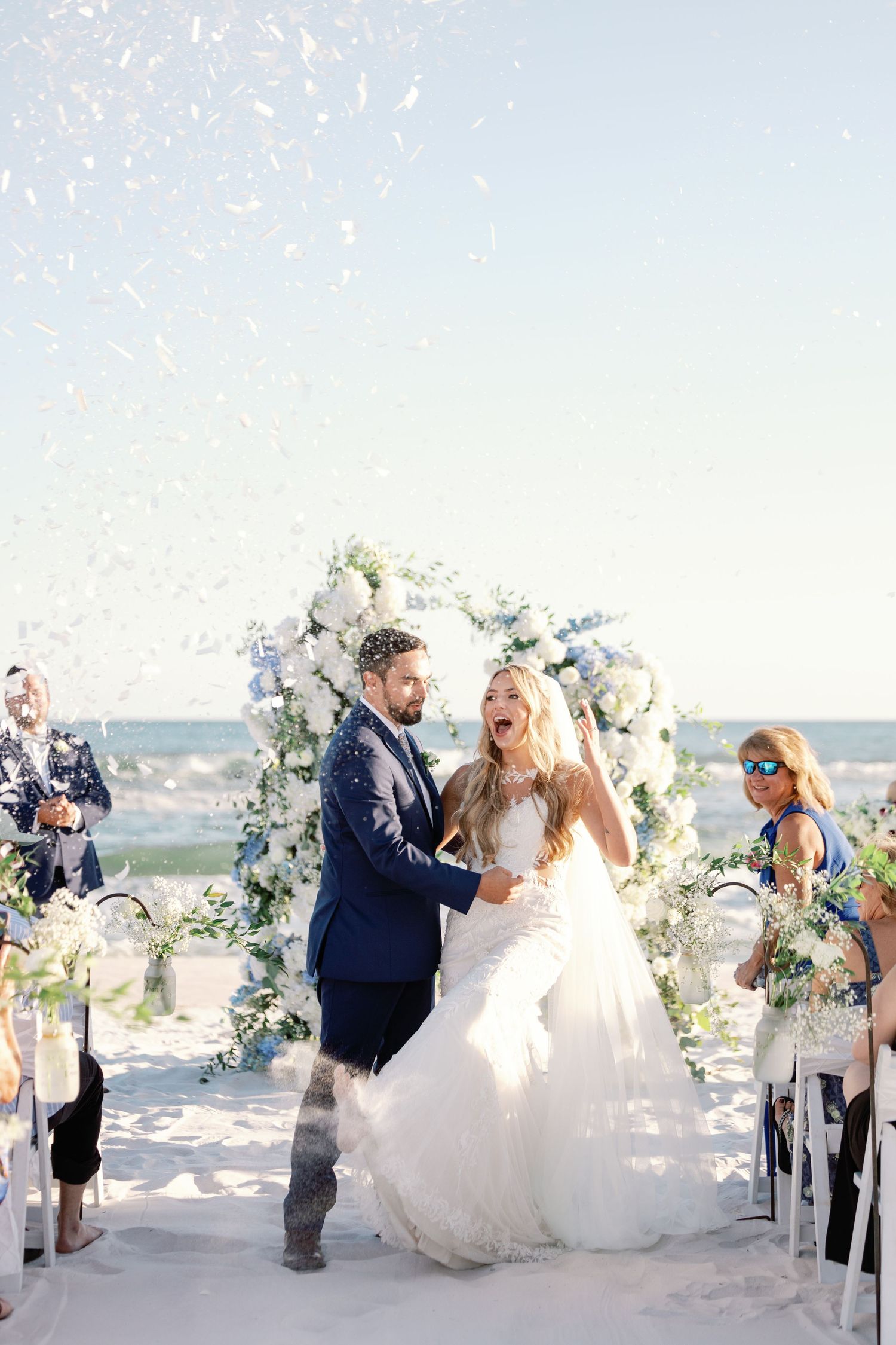 Wedding guests celebrate as couple shares first kiss under floral arch on beach.