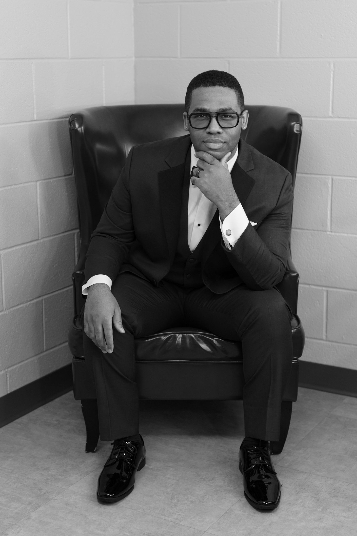 Groom in formal suit and glasses sits reflectively in black leather chair.