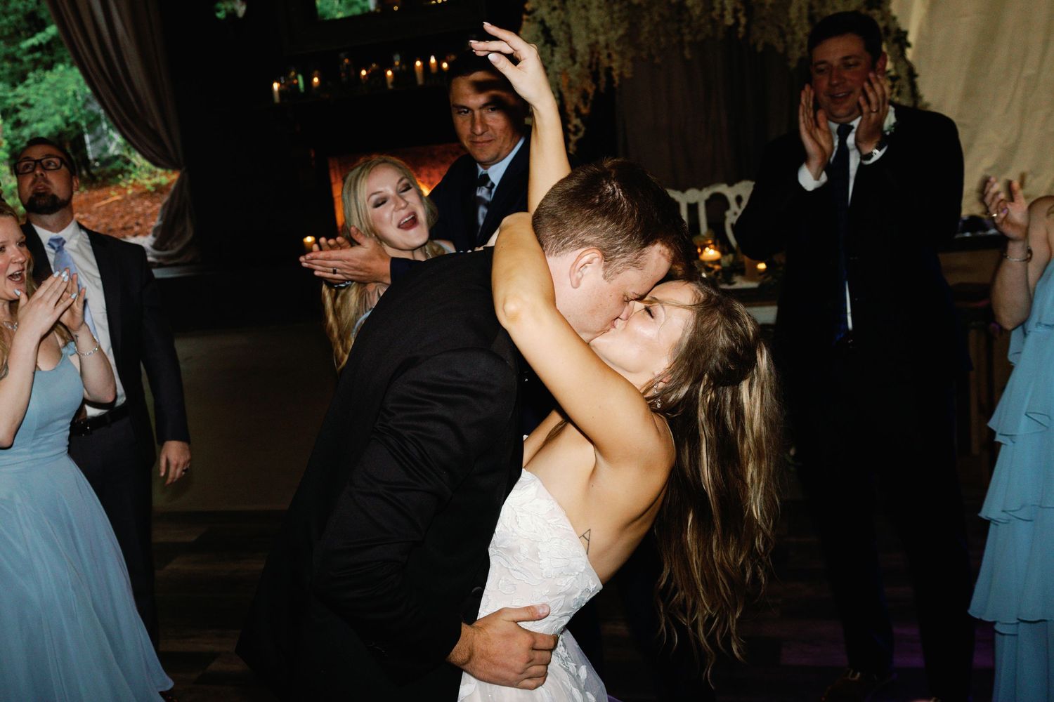 Wedding couple dances under dim lighting surrounded by celebrating guests.