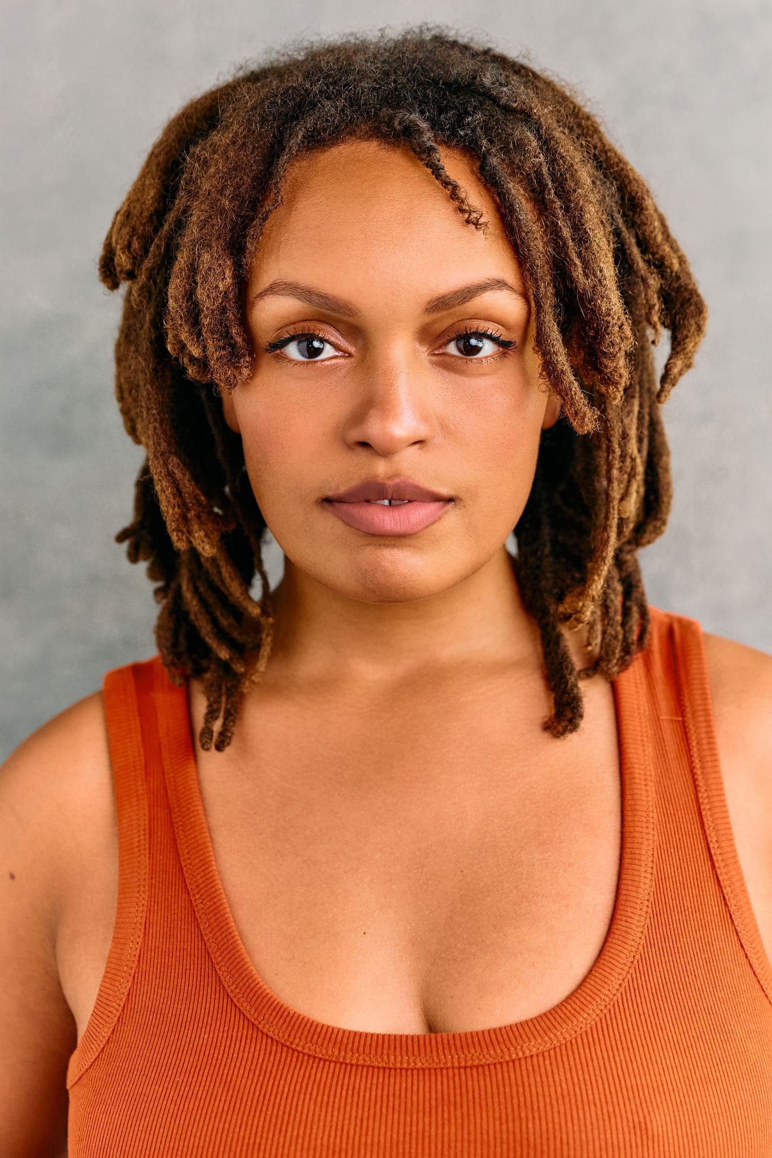 Young woman with brown locs wearing an orange tank top poses against a gray background.