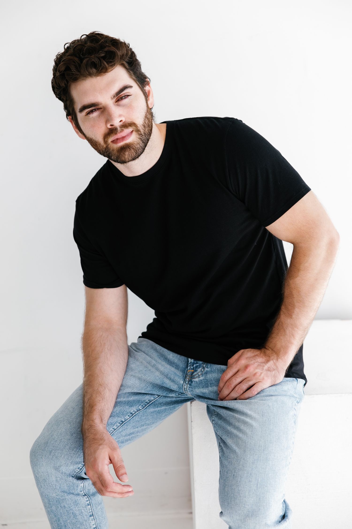 Man in black t-shirt and light jeans poses casually in studio setting.