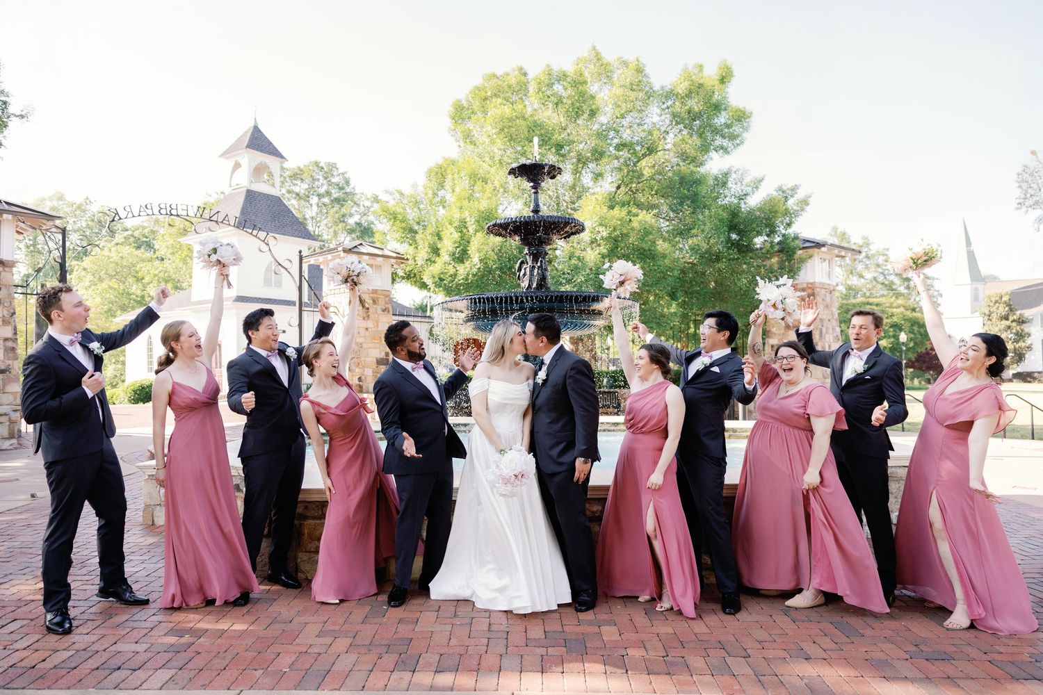Bridal party in burgundy dresses and groomsmen in black suits pose for outdoor portraits.