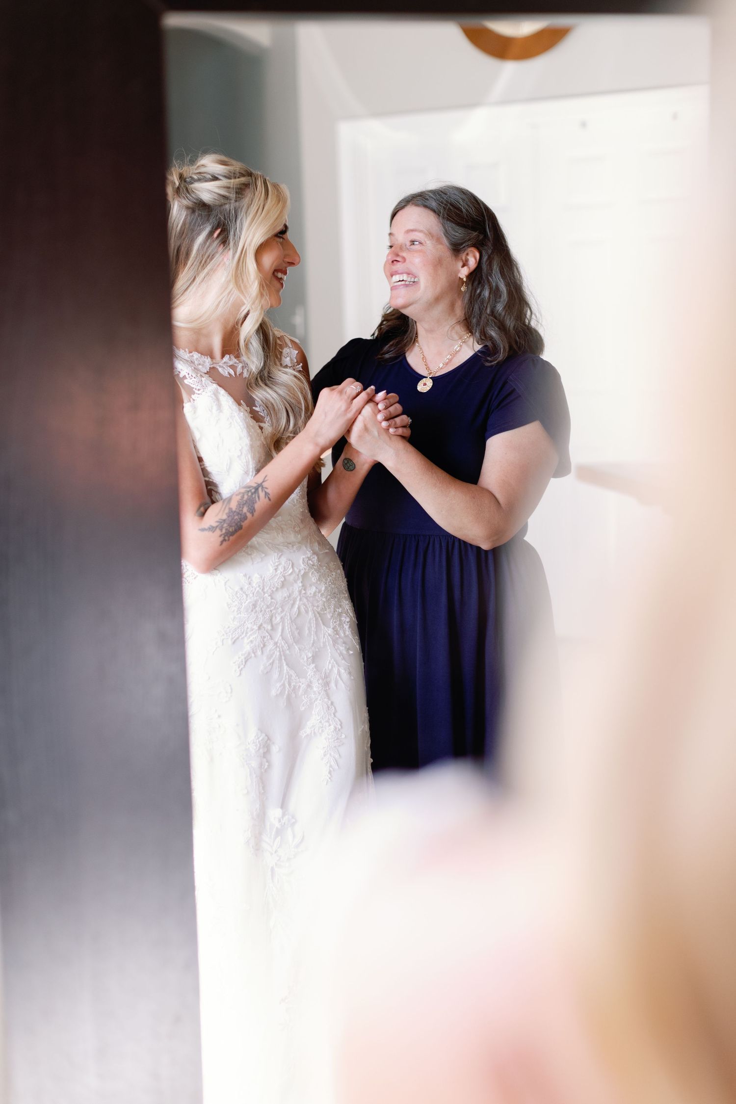 Two women share a joyful moment wearing white and navy dresses in a bridal preparation scene.