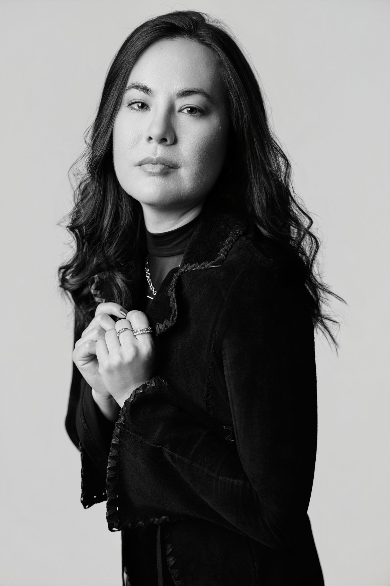 Black and white portrait of a woman with long dark hair wearing a black outfit, hands clasped, studio background.