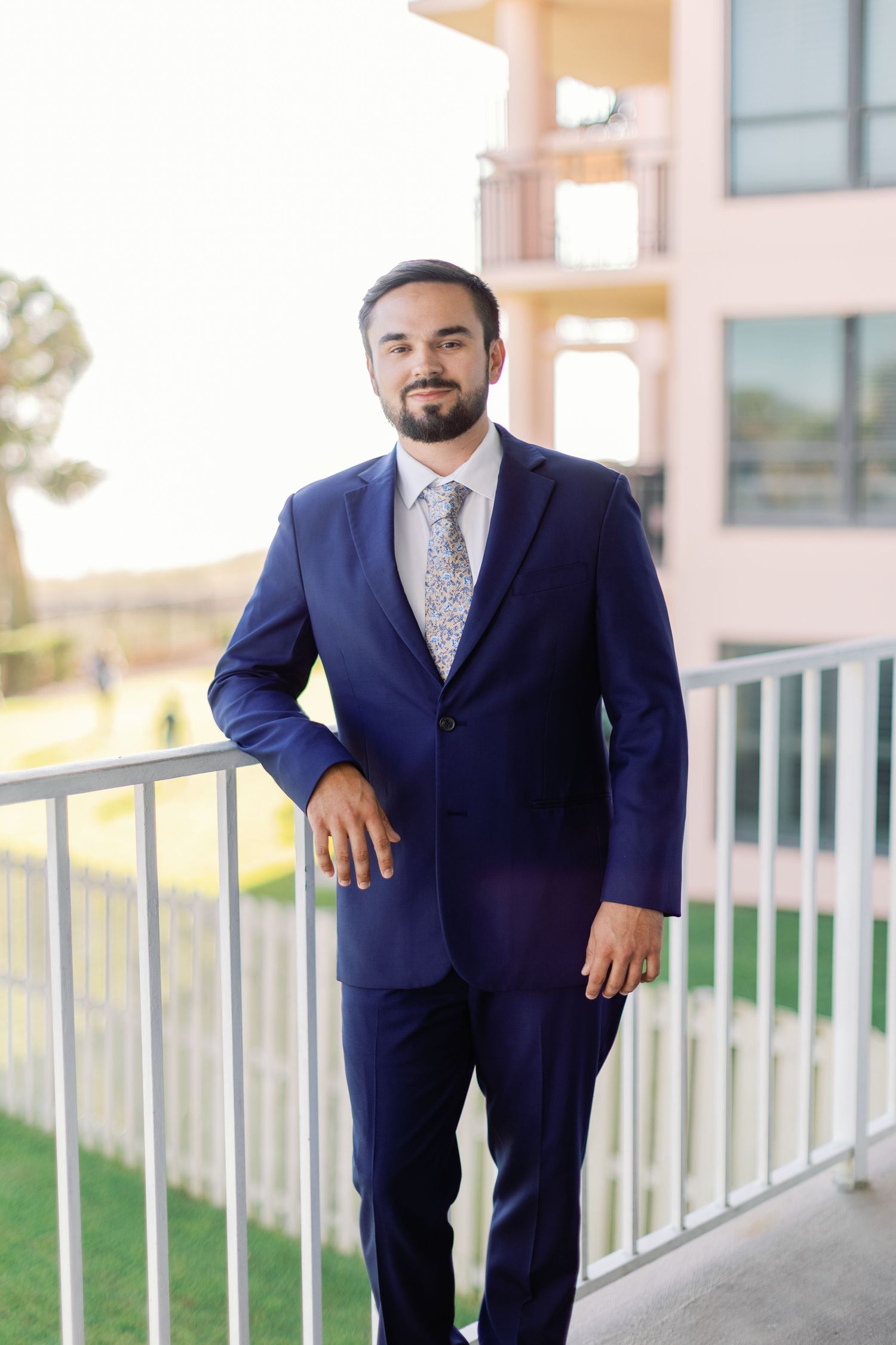 A groom poses confidently on a balcony wearing a fitted navy suit and floral tie.