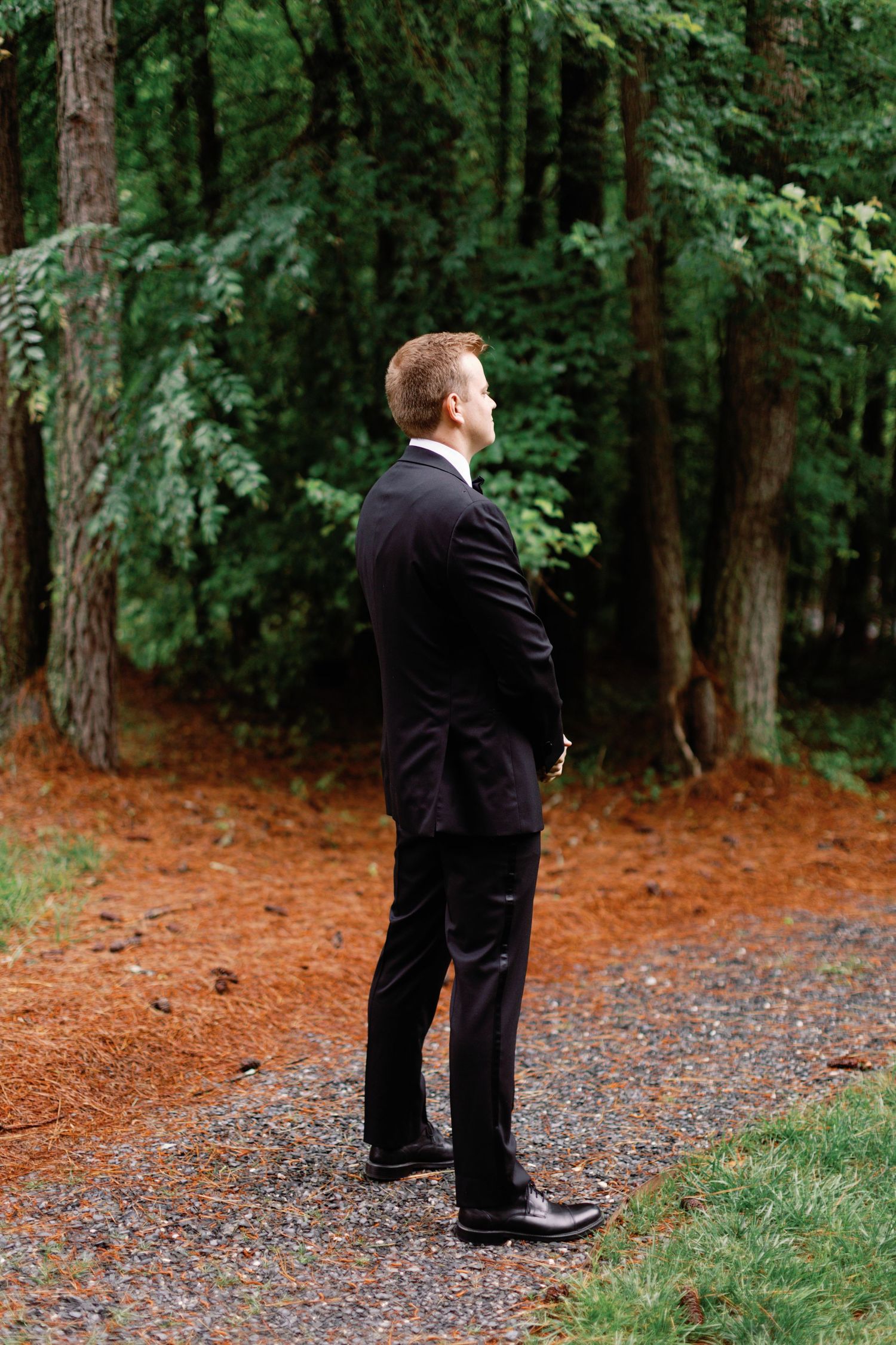 Person in black suit stands contemplatively on a forest path surrounded by pine trees.