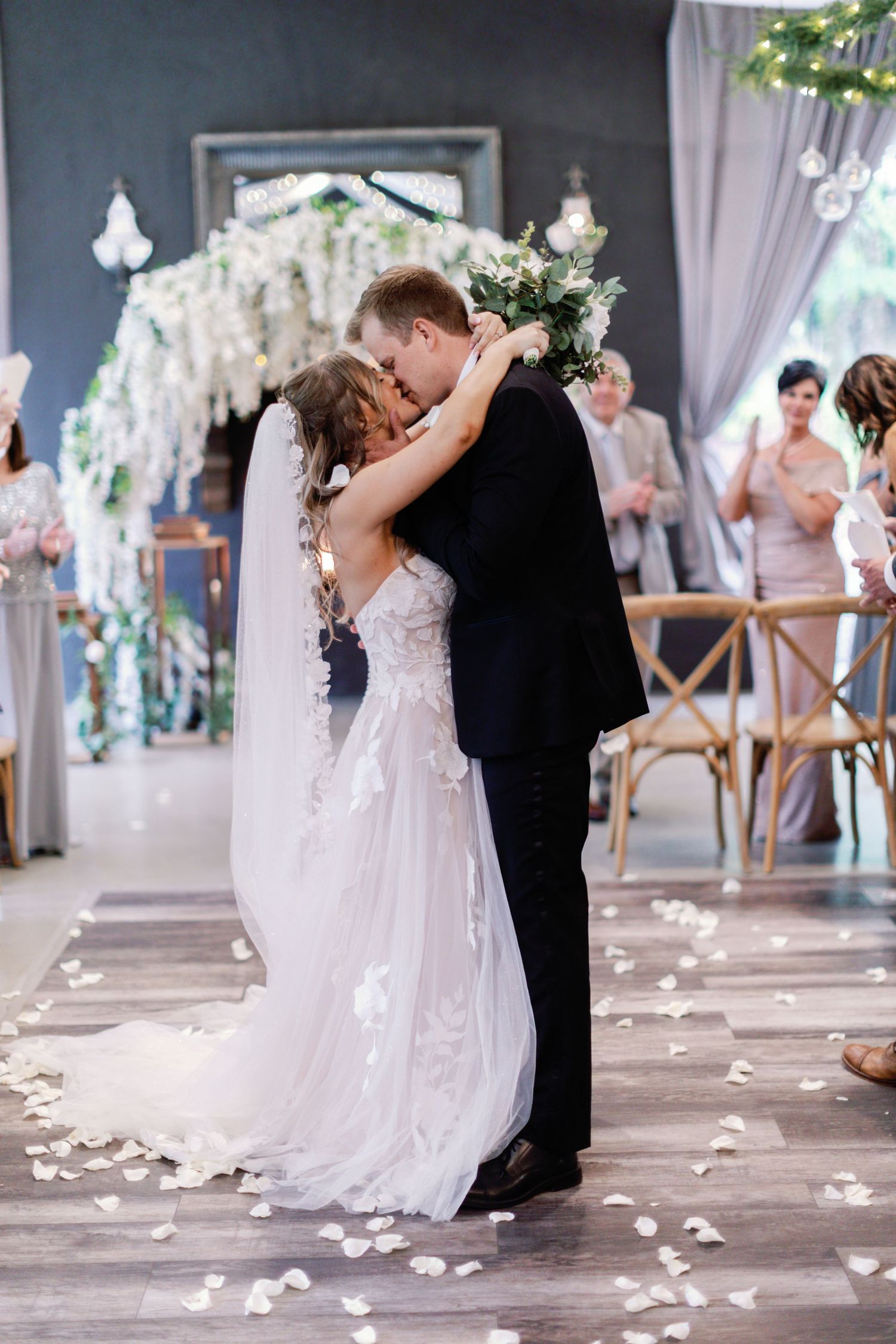 Romantic wedding moments captured under cascading white flowers with rose petals scattered on the floor.