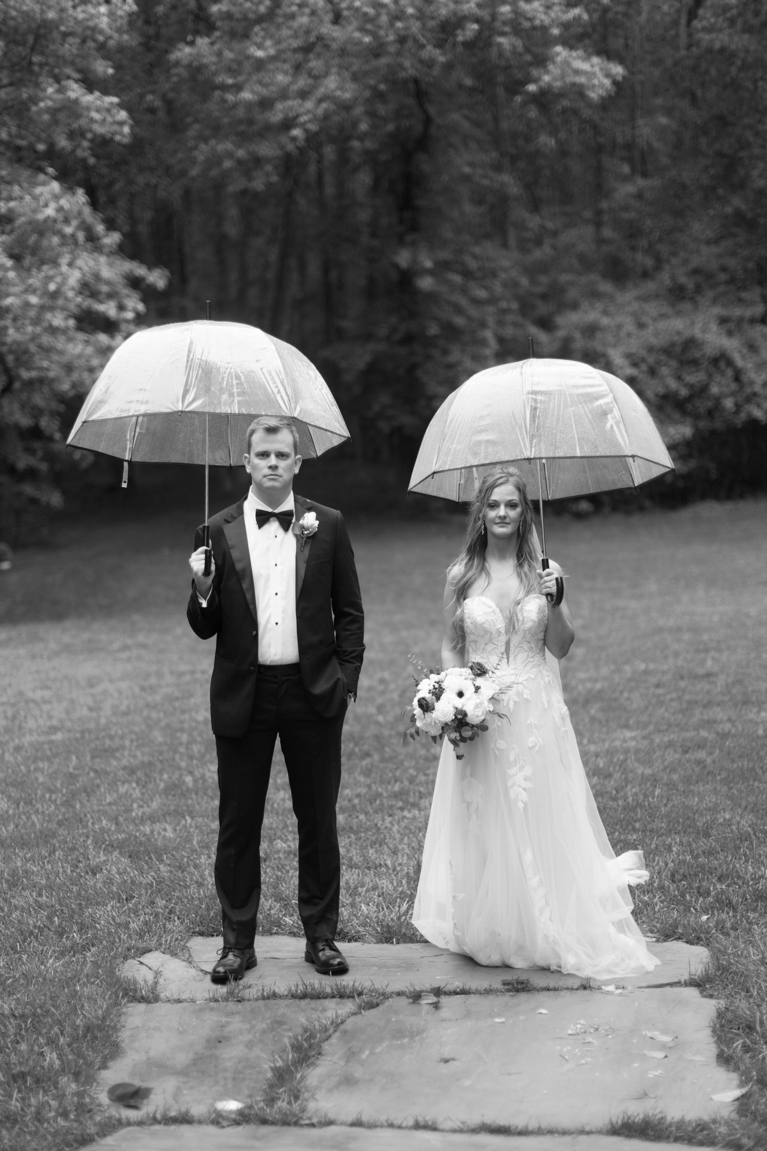 Couple in wedding attire standing with clear umbrellas on rainy day.