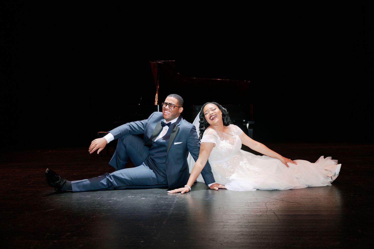 Newlyweds in wedding attire sitting playfully on a dark stage floor.