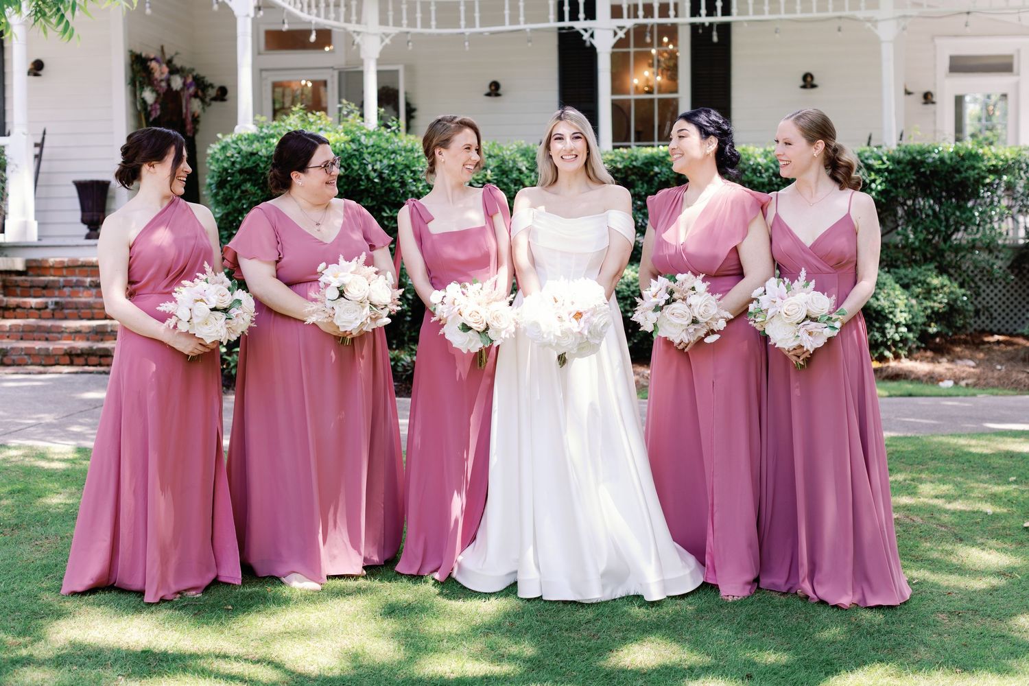 Bridal party in dusty rose dresses arranged in a line on a garden lawn.