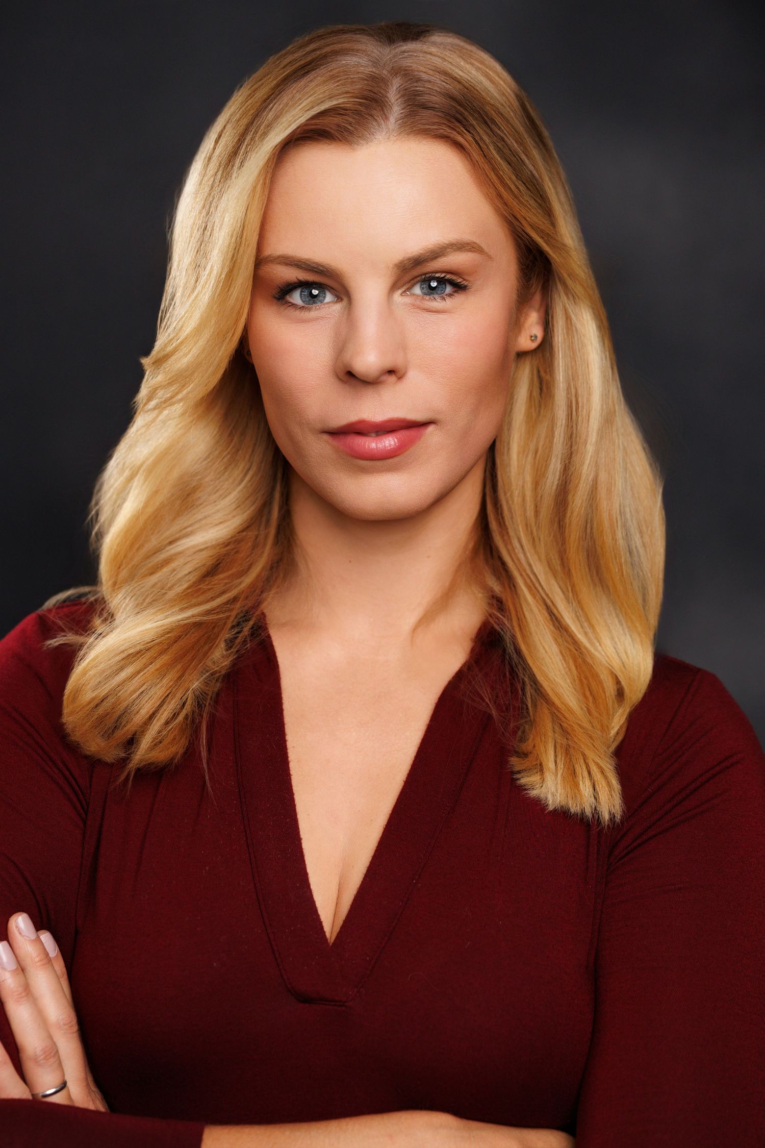 Professional headshot of someone with blonde wavy hair wearing a burgundy top against a dark background.