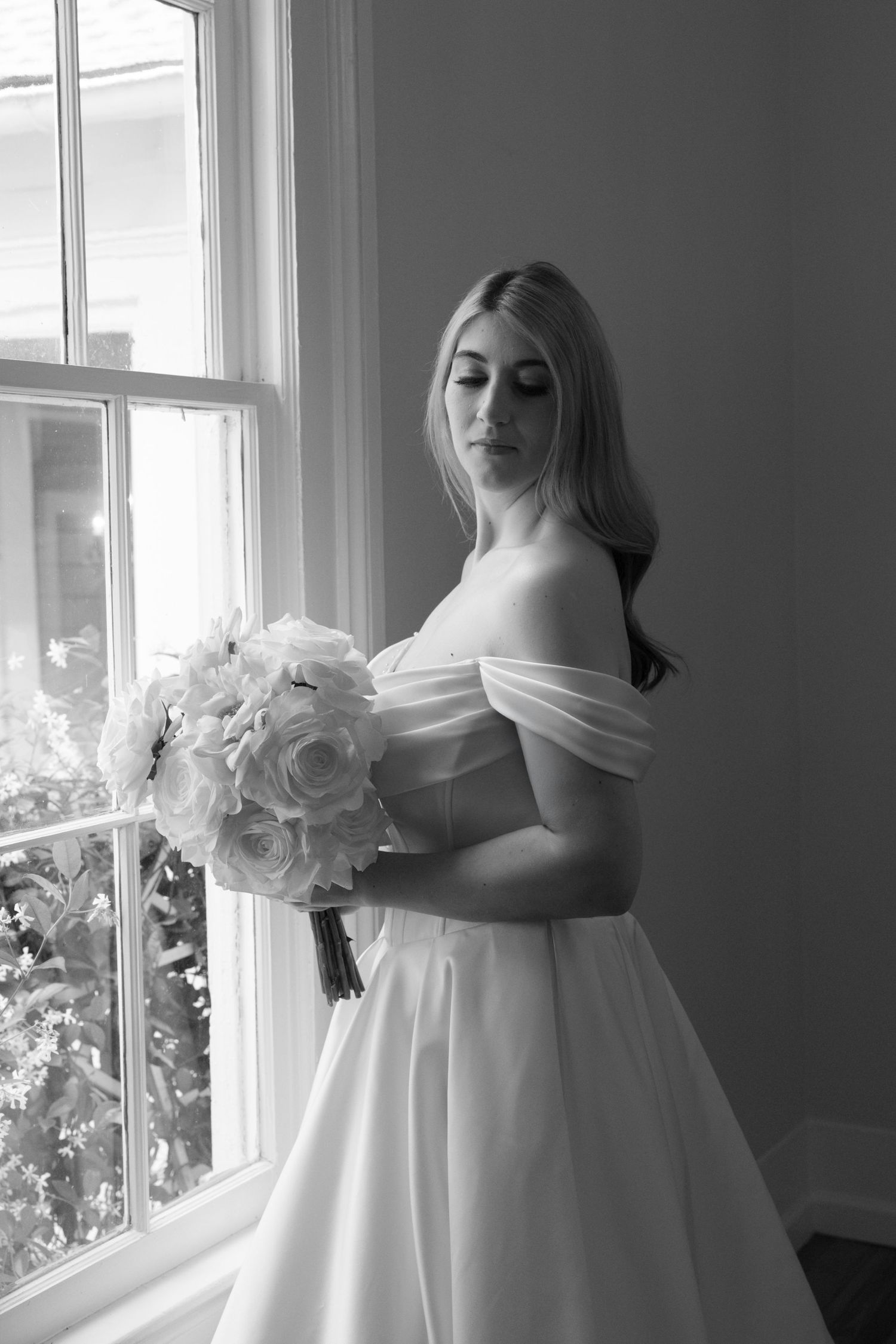 Elegant black and white portrait of someone in a white bridal gown with floral details near a window.