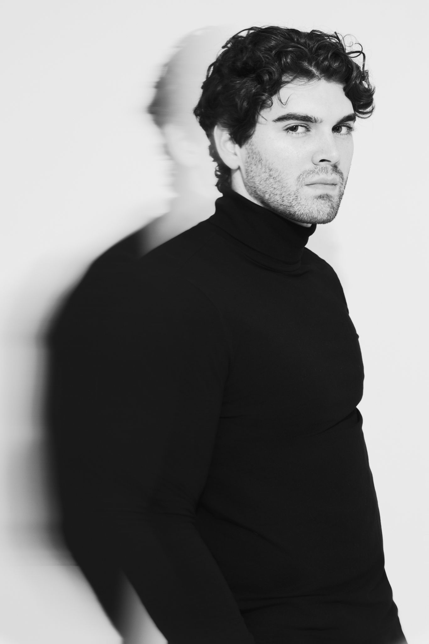 A black and white portrait of a person in a black turtleneck sweater with dark curly hair against a light background.