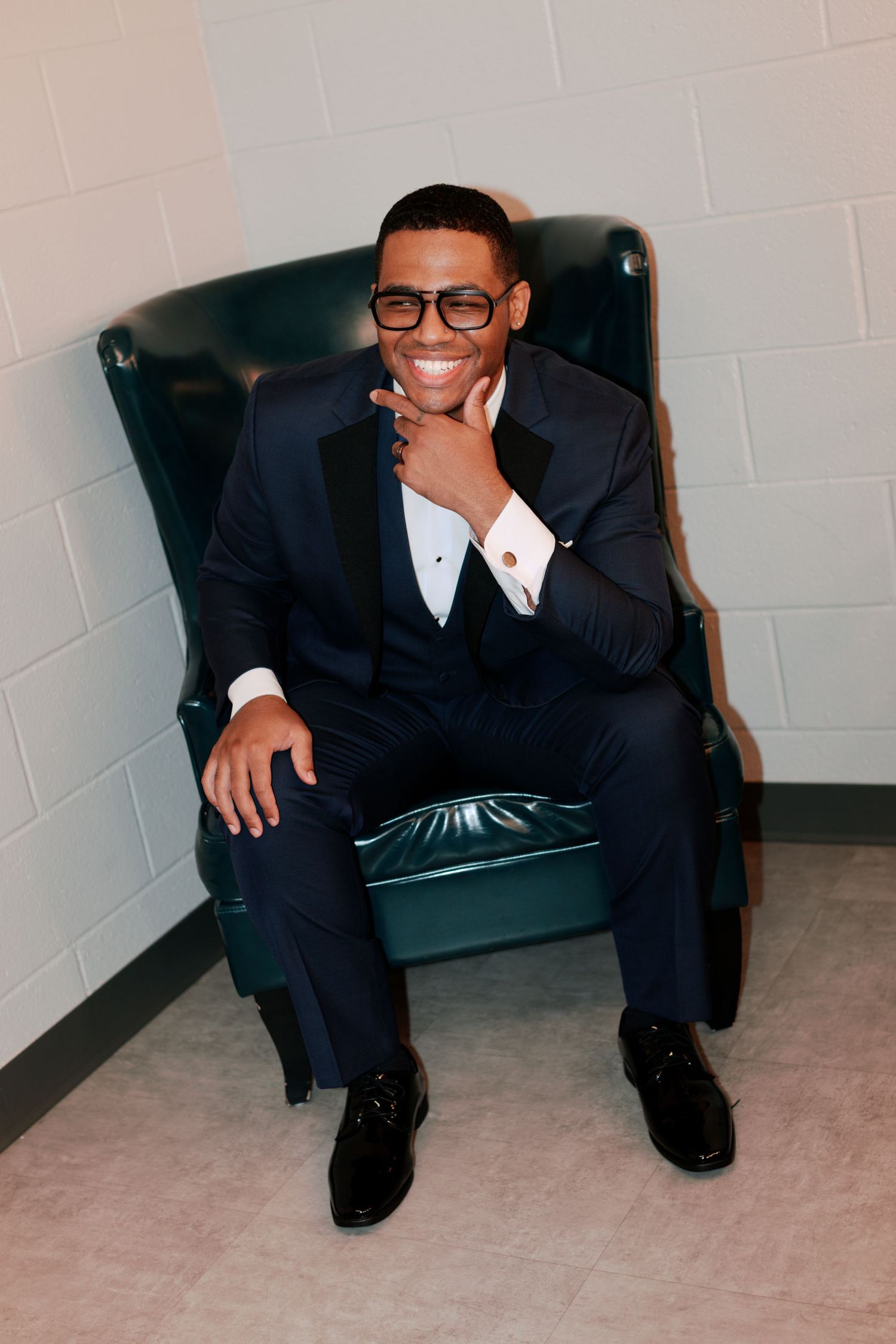 Groom in tuxedo and glasses relaxes in leather chair while getting ready.