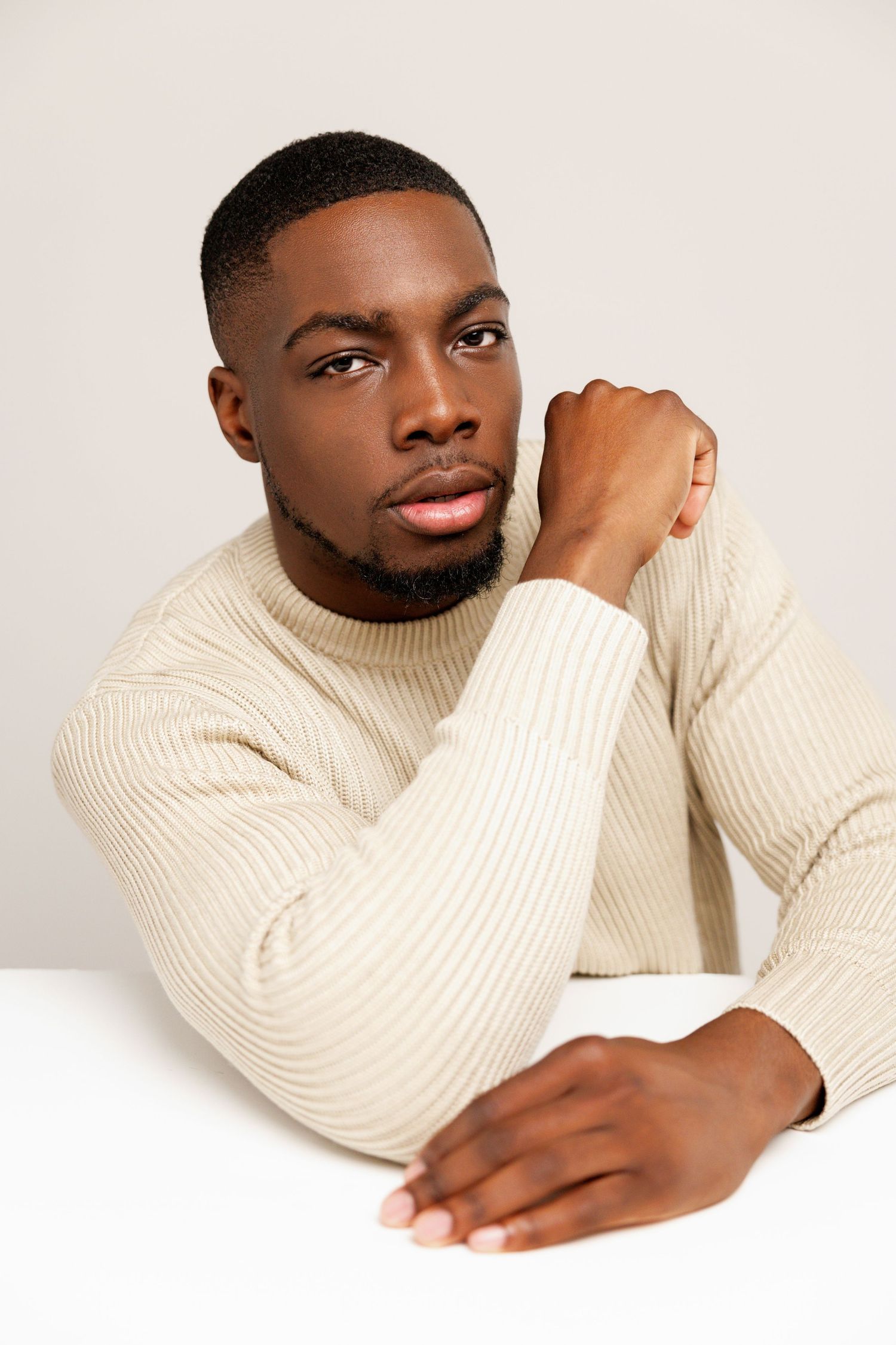 A portrait of a person in a cream ribbed sweater posing thoughtfully against a white backdrop.