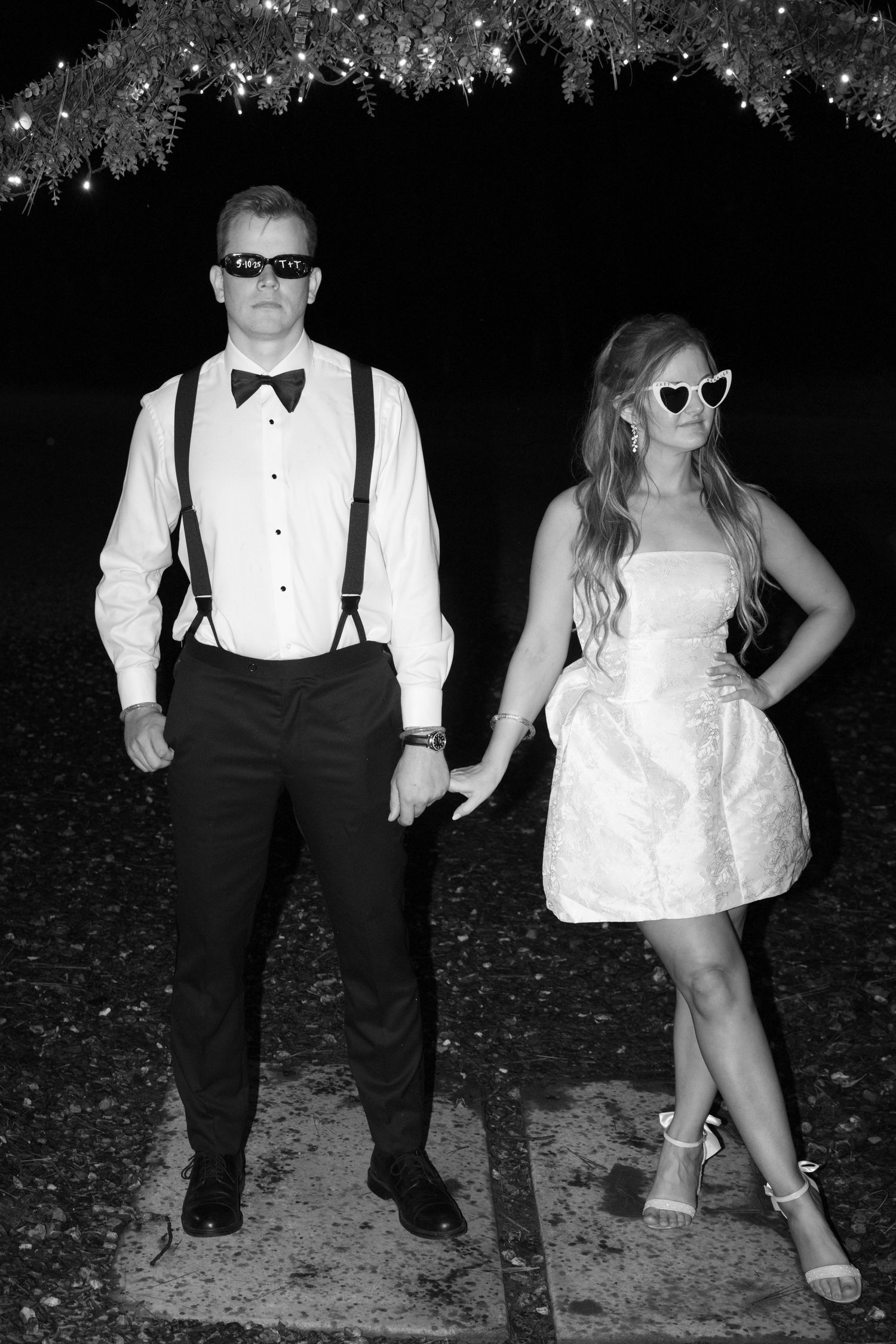 A stylishly dressed couple in sunglasses poses under twinkling lights at night.