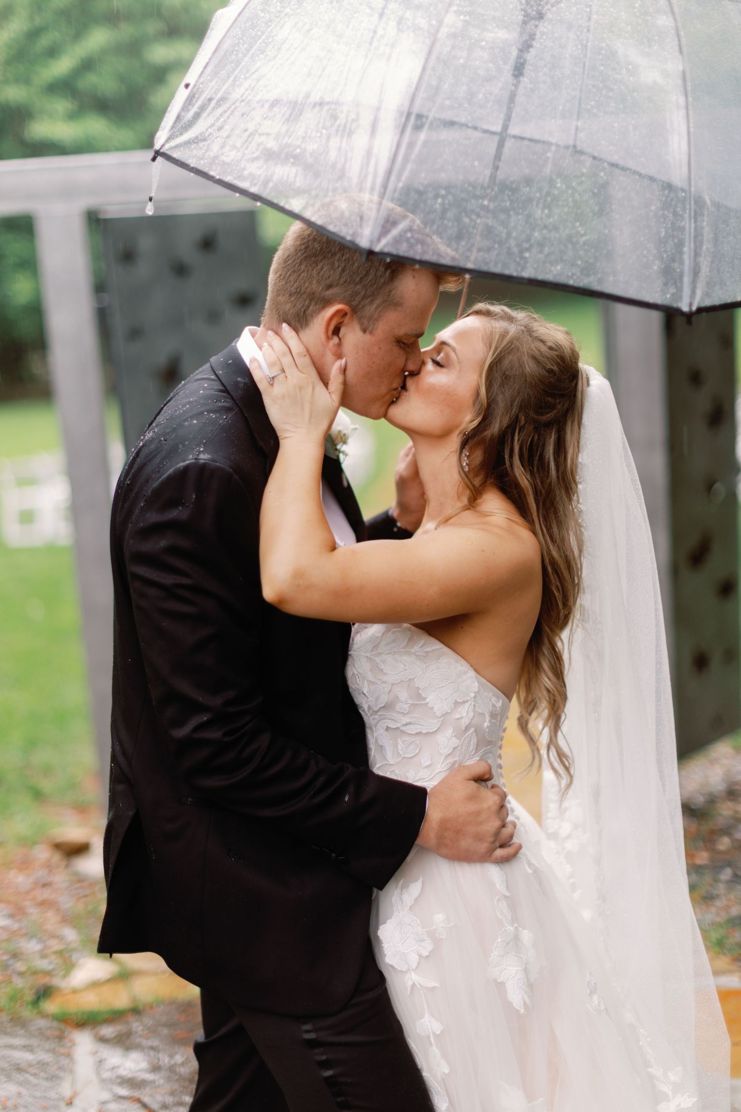 Newlyweds share a kiss beneath a transparent umbrella on their rainy wedding day.