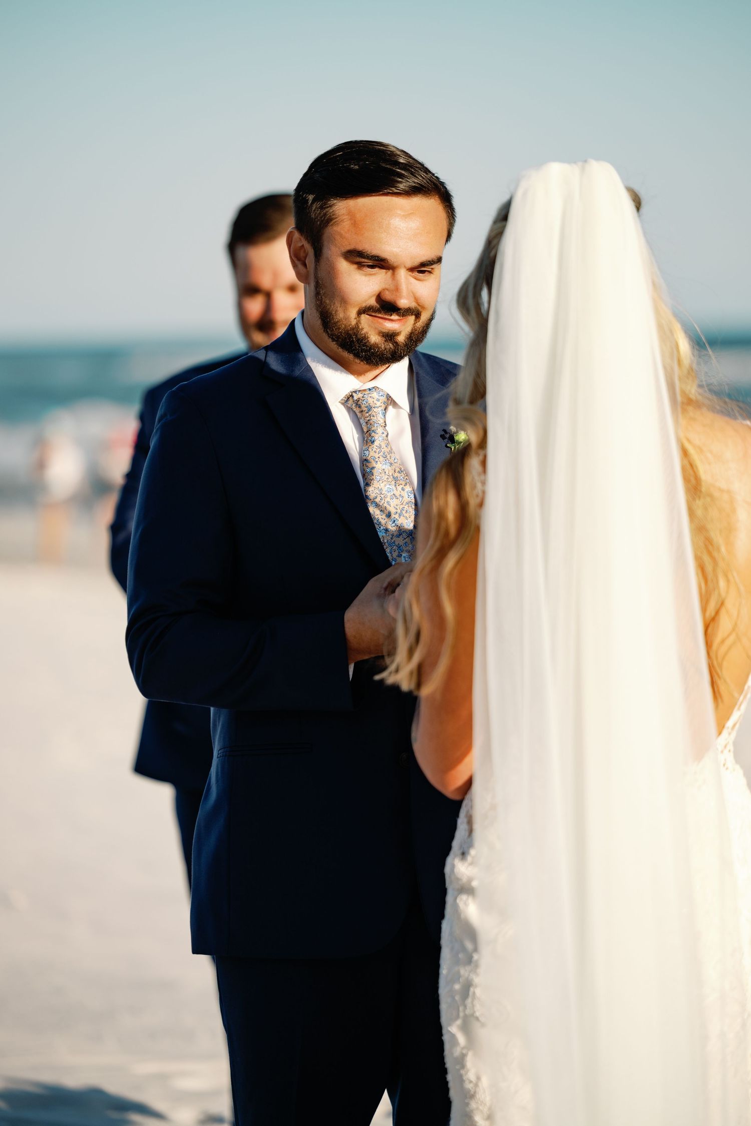 Romantic beach wedding photoshoot with couple embracing against blue sky backdrop.