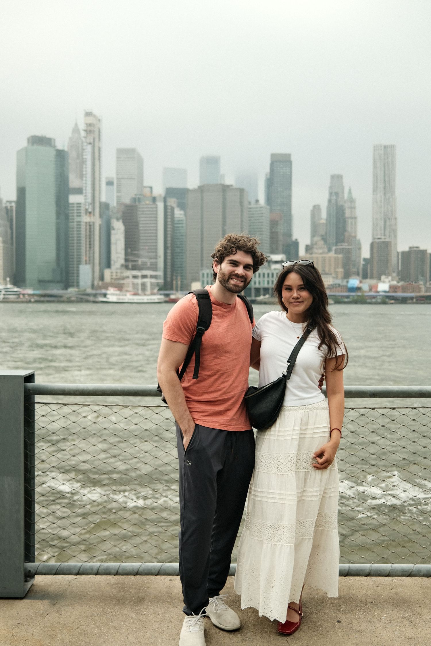 Couple posing along waterfront with Manhattan skyline visible across the river.