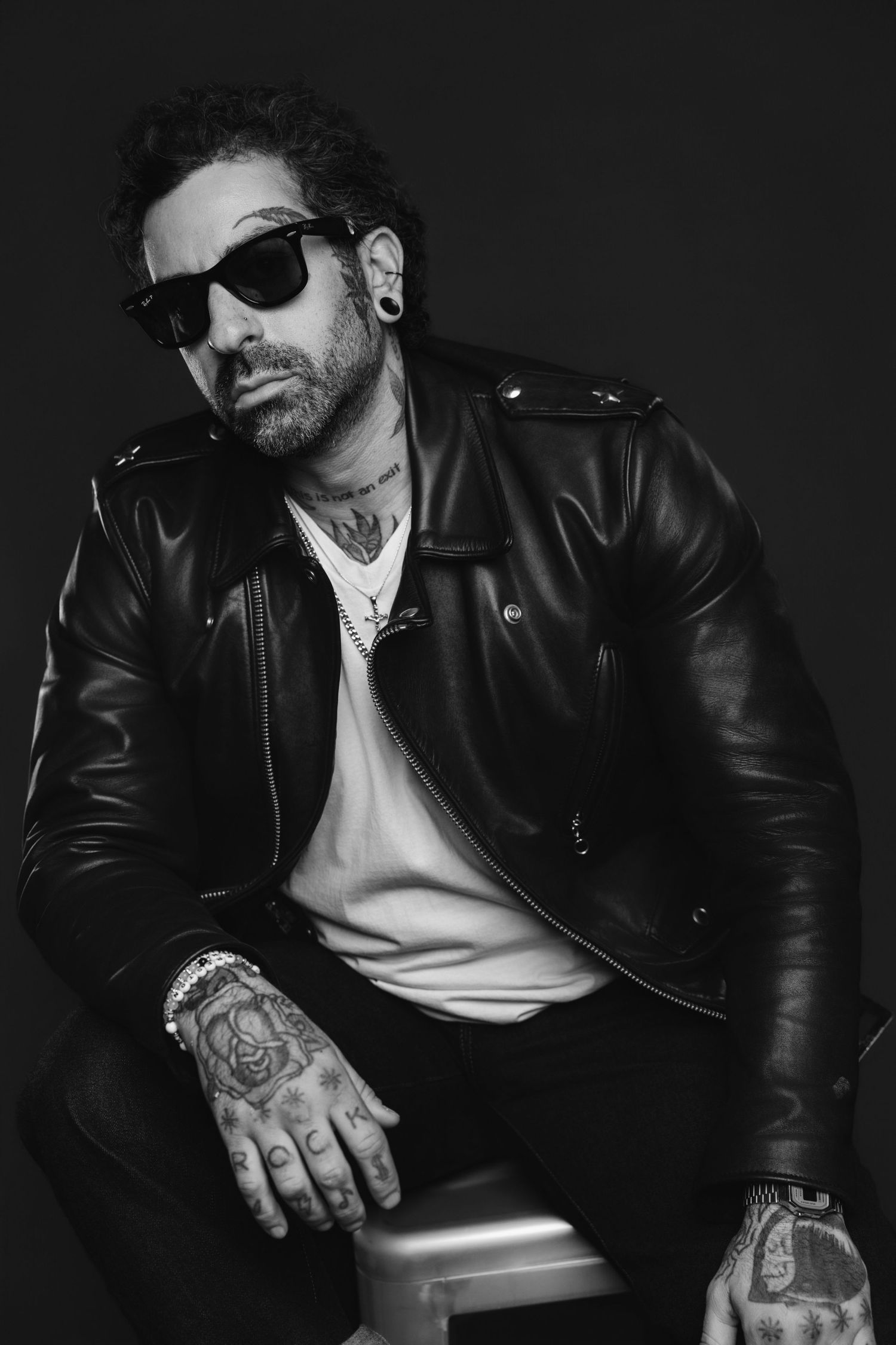Black and white portrait of person in leather jacket and sunglasses showing tattoos while seated against dark background.