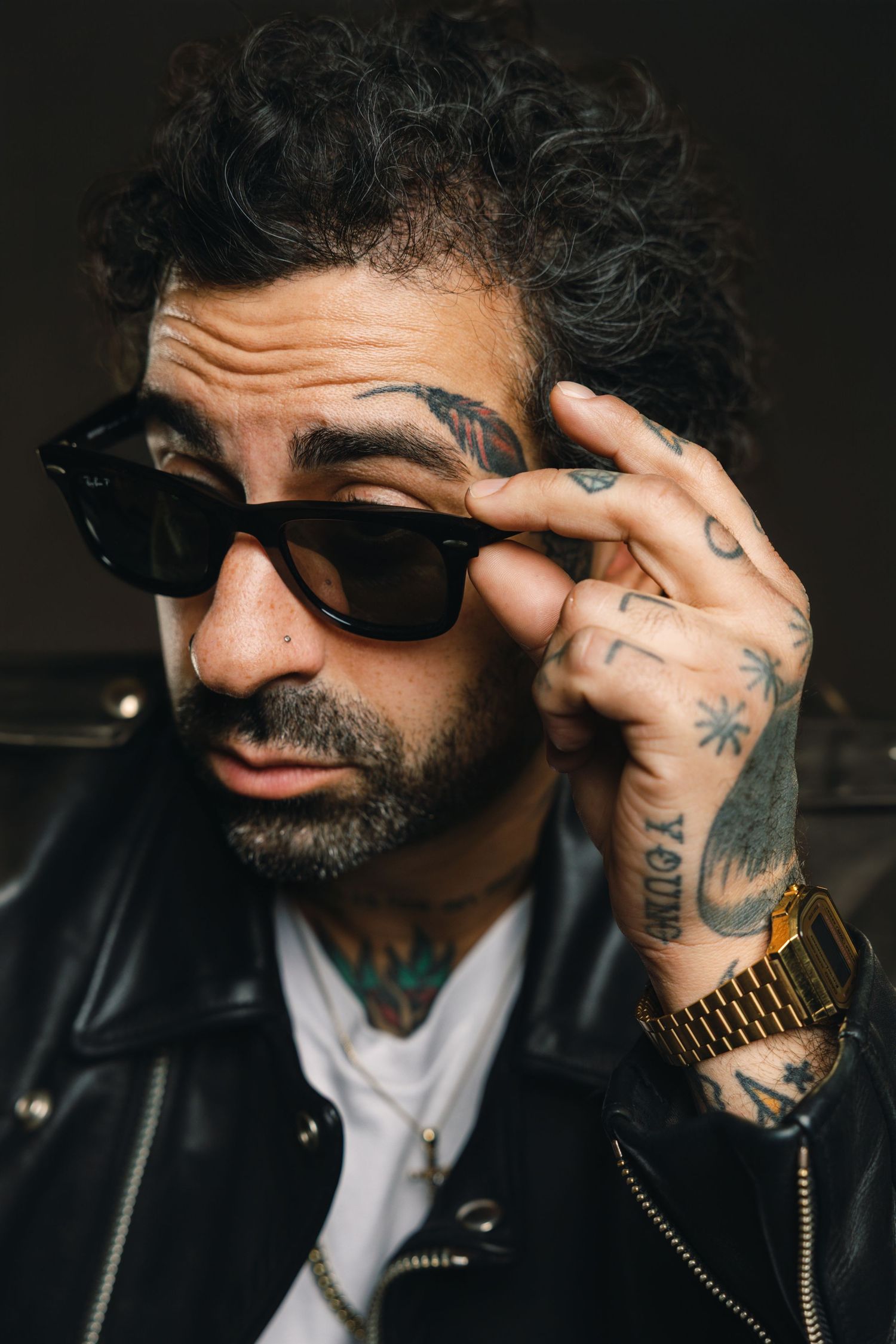 Close-up portrait of someone in sunglasses and leather jacket with visible tattoos and gold watch adjusting their frames.