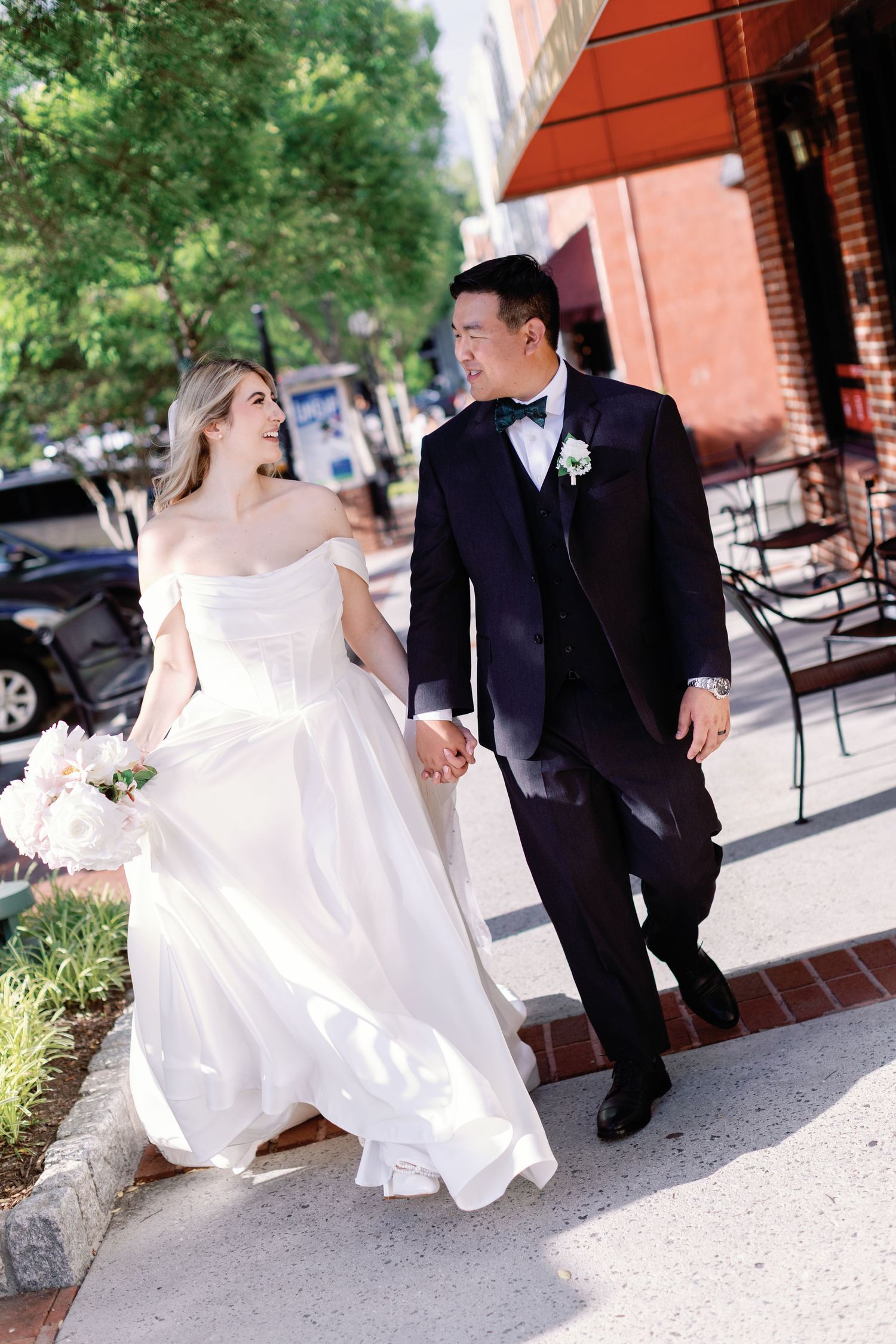 Couple in wedding attire walking together on sidewalk under red awning.