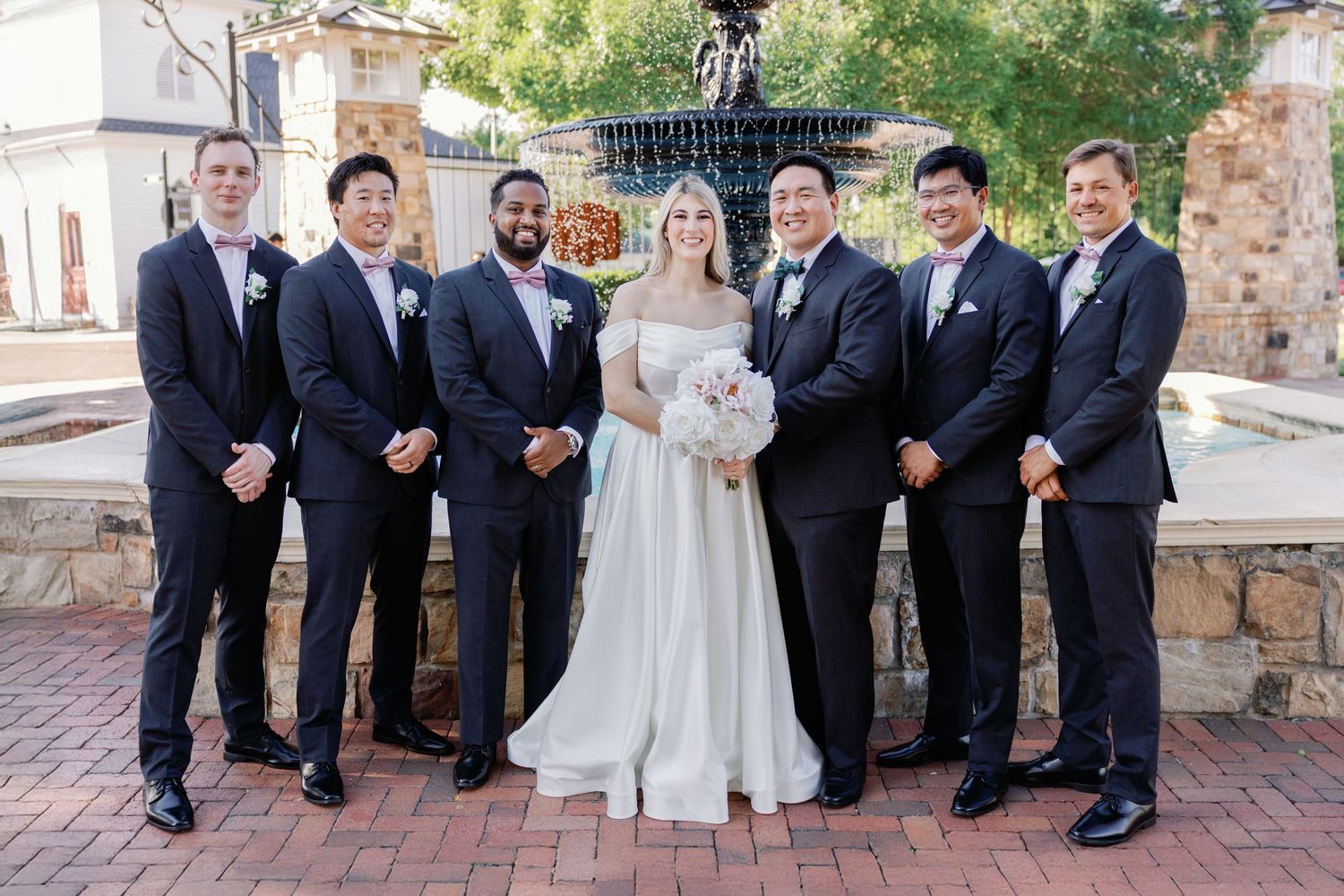 Groomsmen in dark suits pose with couple on brick pathway during wedding portraits.