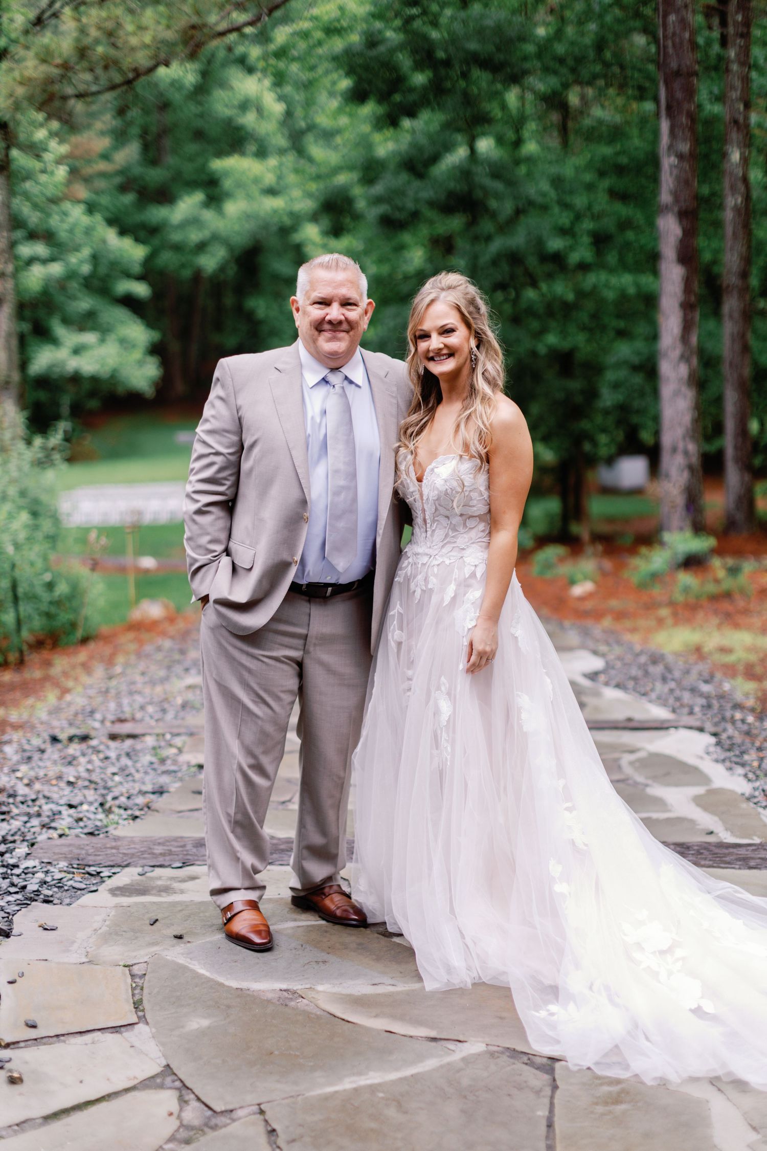 Two people pose together on a stone pathway, one in a gray suit and one in a wedding dress.