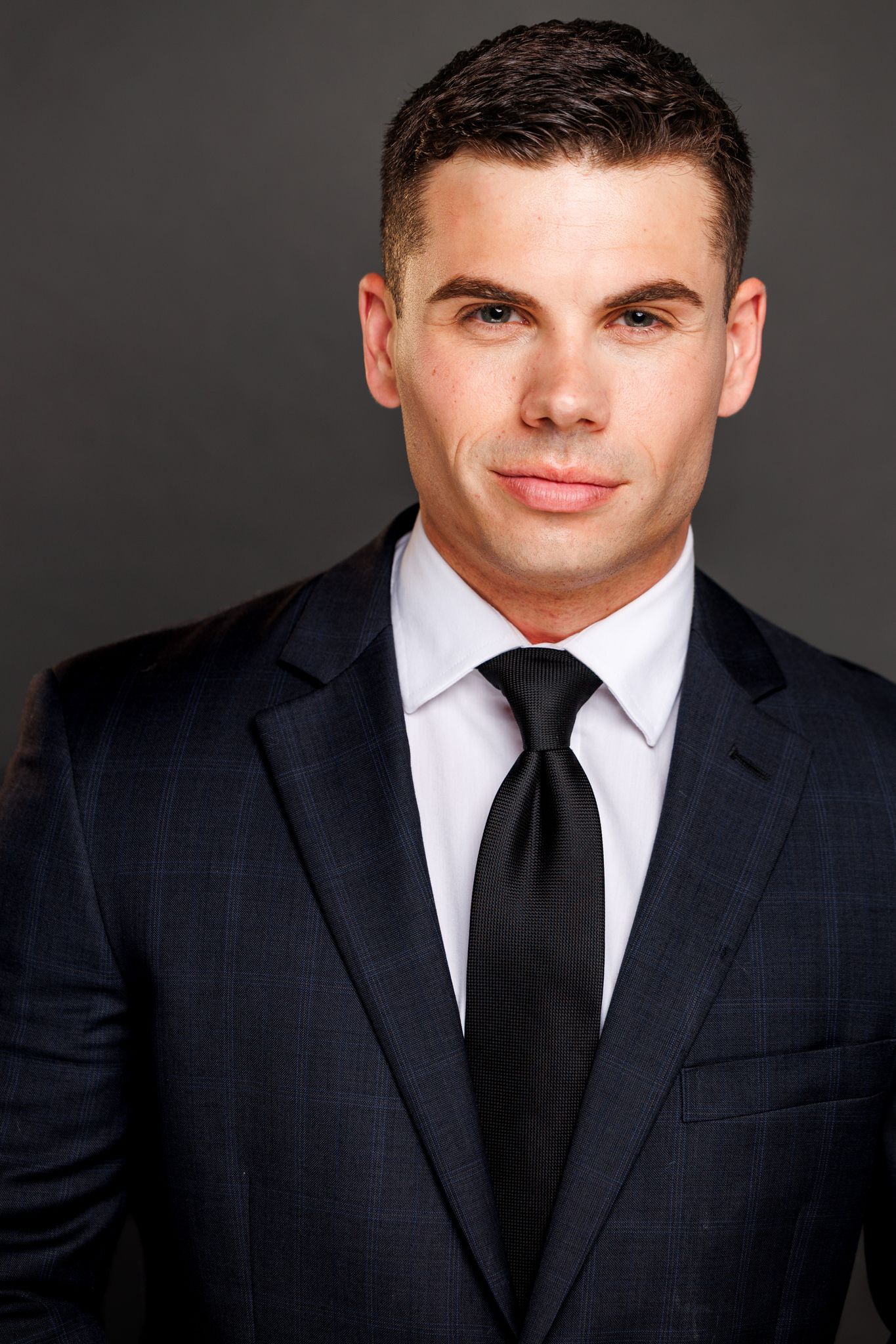 Professional headshot of someone in a dark suit and tie against a grey background.