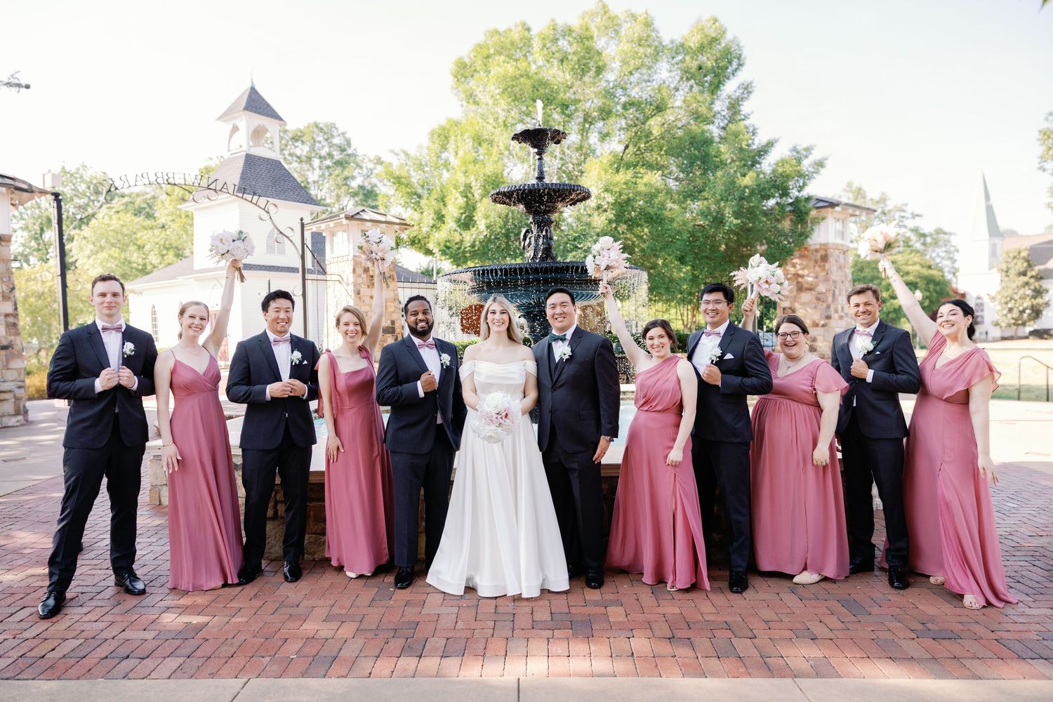 Large wedding party in burgundy and black attire poses for formal group photos outdoors.