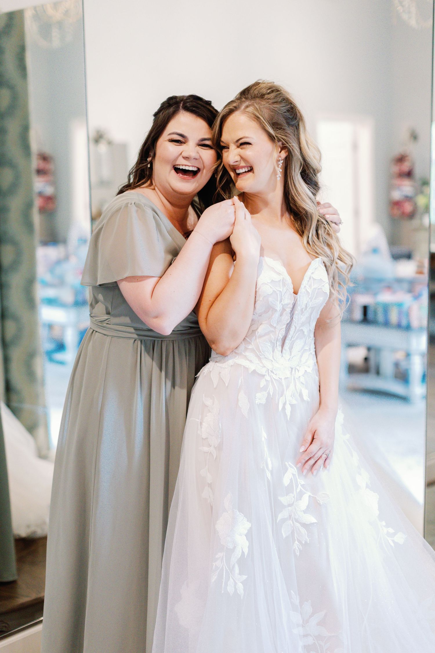 Two women share a joyful moment in a bridal boutique, one wearing a gray dress and one in a white wedding gown.