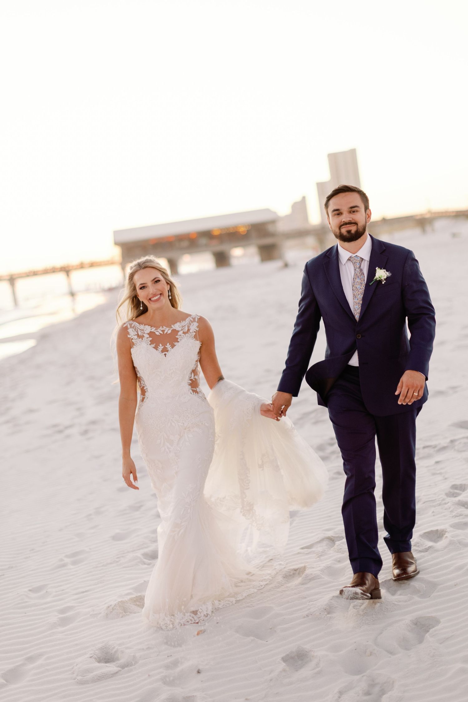 Wedding couple walks hand in hand along pristine beach dunes in elegant attire.