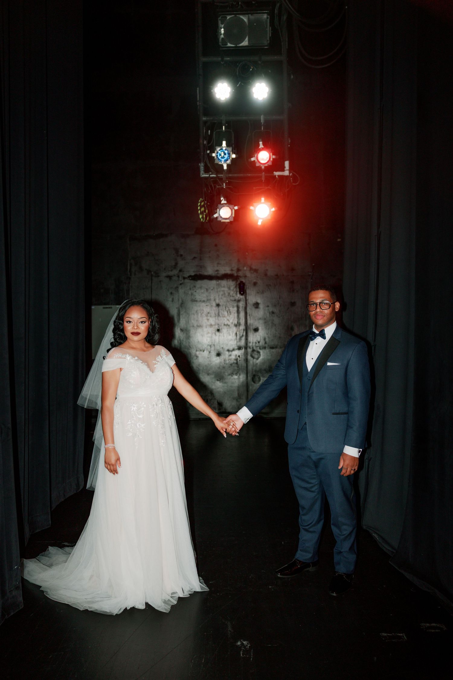 Elegant wedding couple posing beneath theatrical lighting in a dark industrial space.