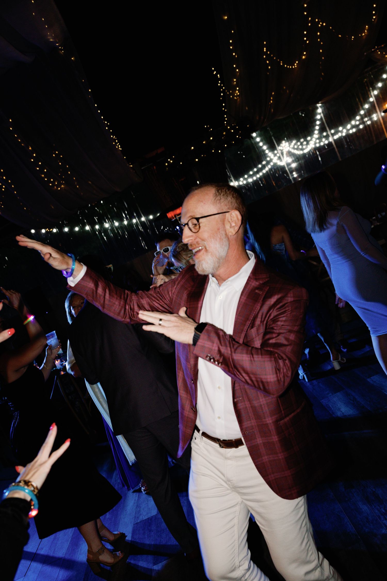A person in a burgundy blazer dances energetically under twinkling string lights at a party.