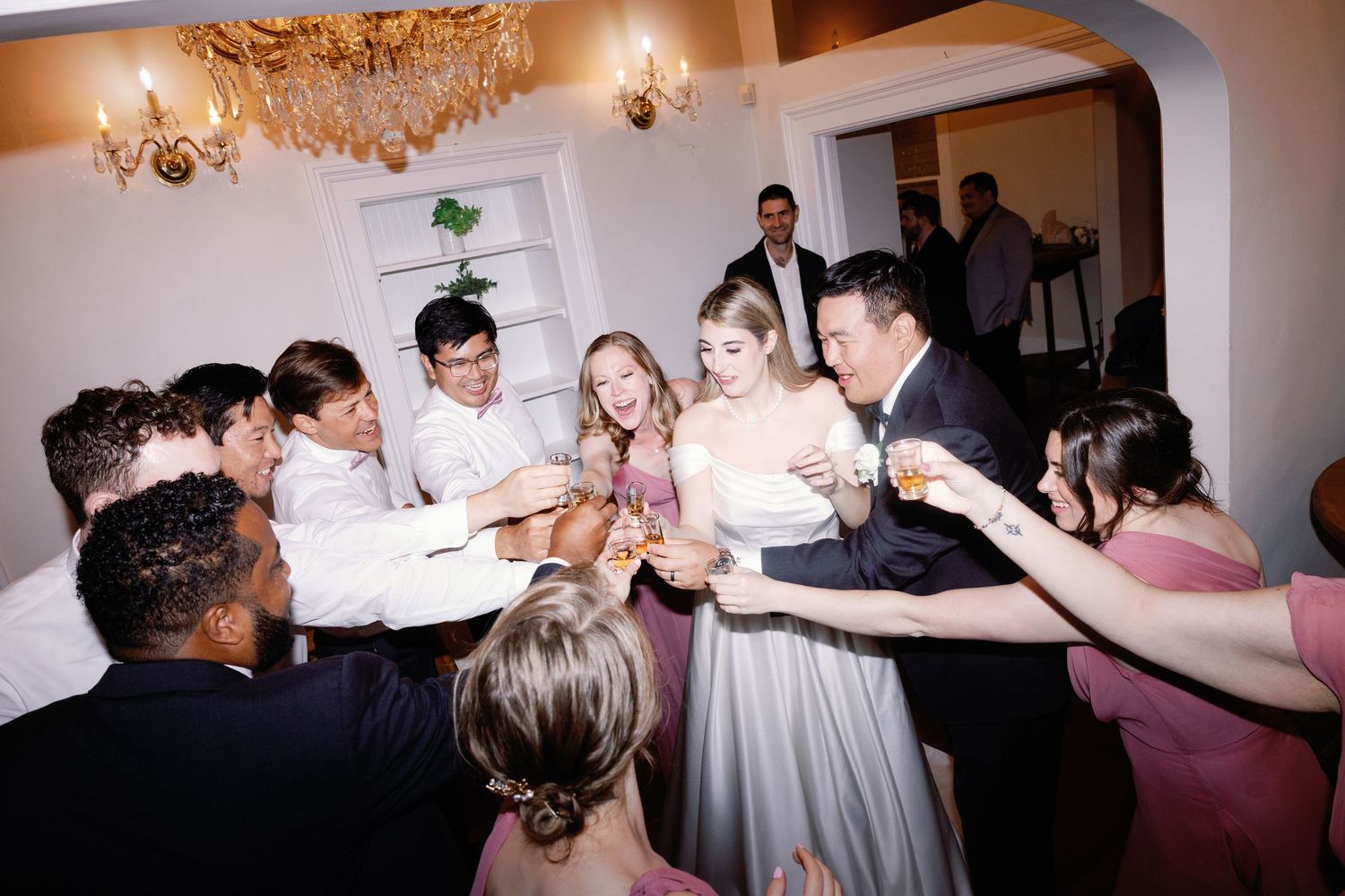 Guests dancing together at a lively wedding reception under chandeliers.