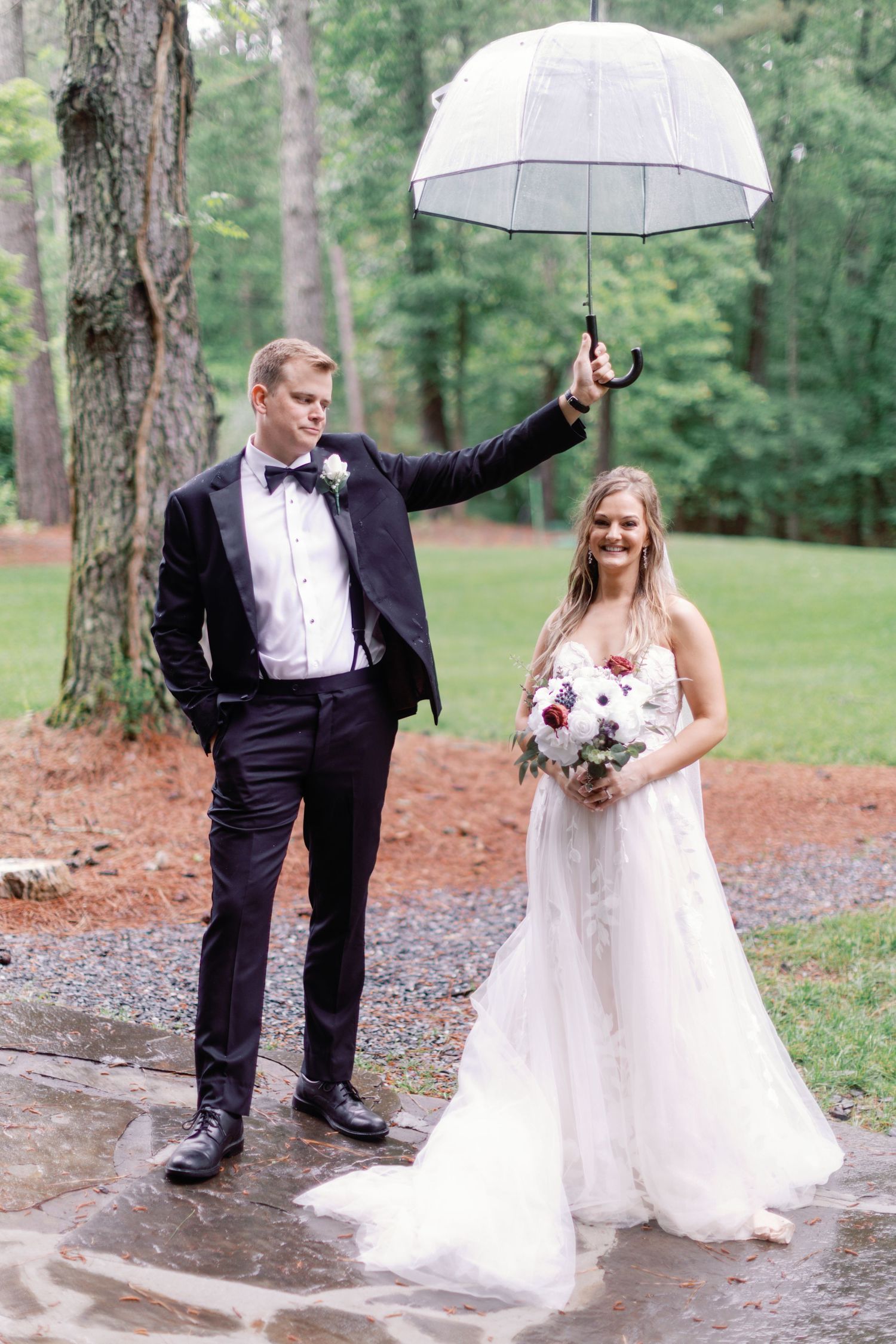 Wedding couple playfully poses with umbrella on wet grounds surrounded by trees.