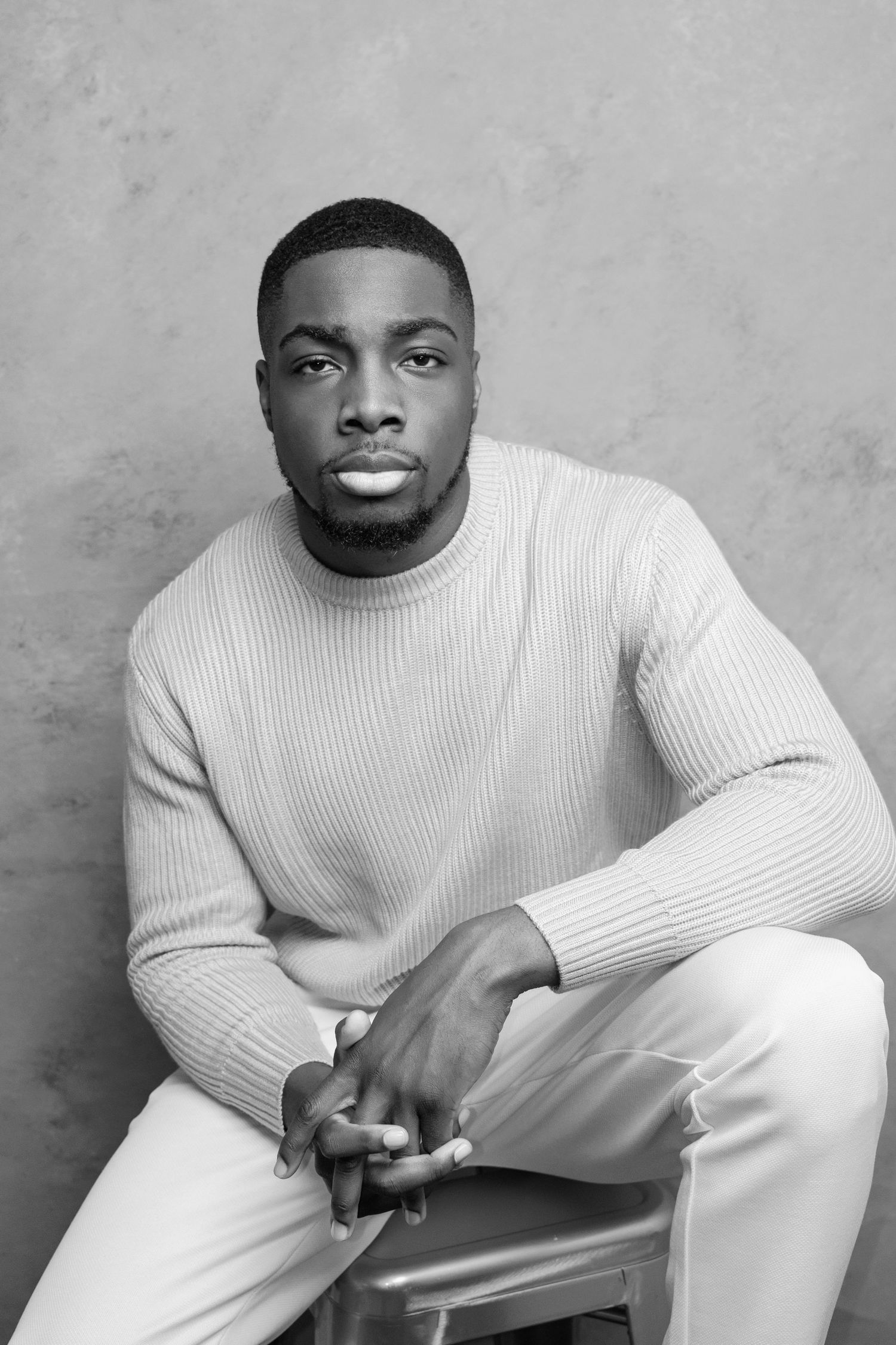 A black and white portrait of someone in a ribbed sweater sitting on a stool.