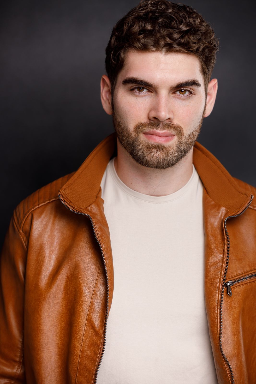 Young man with curly hair and beard wearing a brown leather jacket over a cream shirt against dark background.