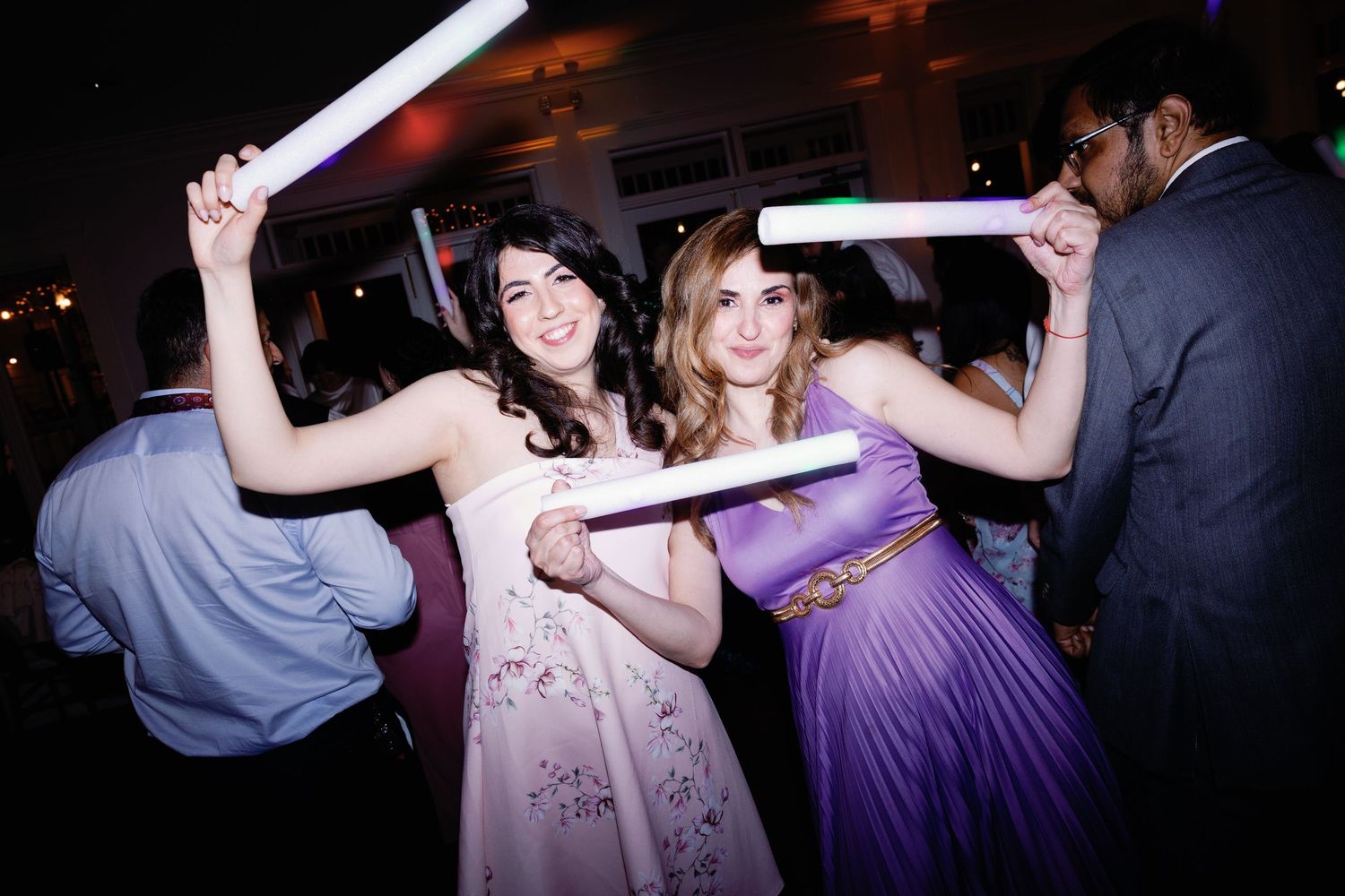 Wedding guests dance with glowing LED foam sticks on a dimly lit dance floor at a reception party.