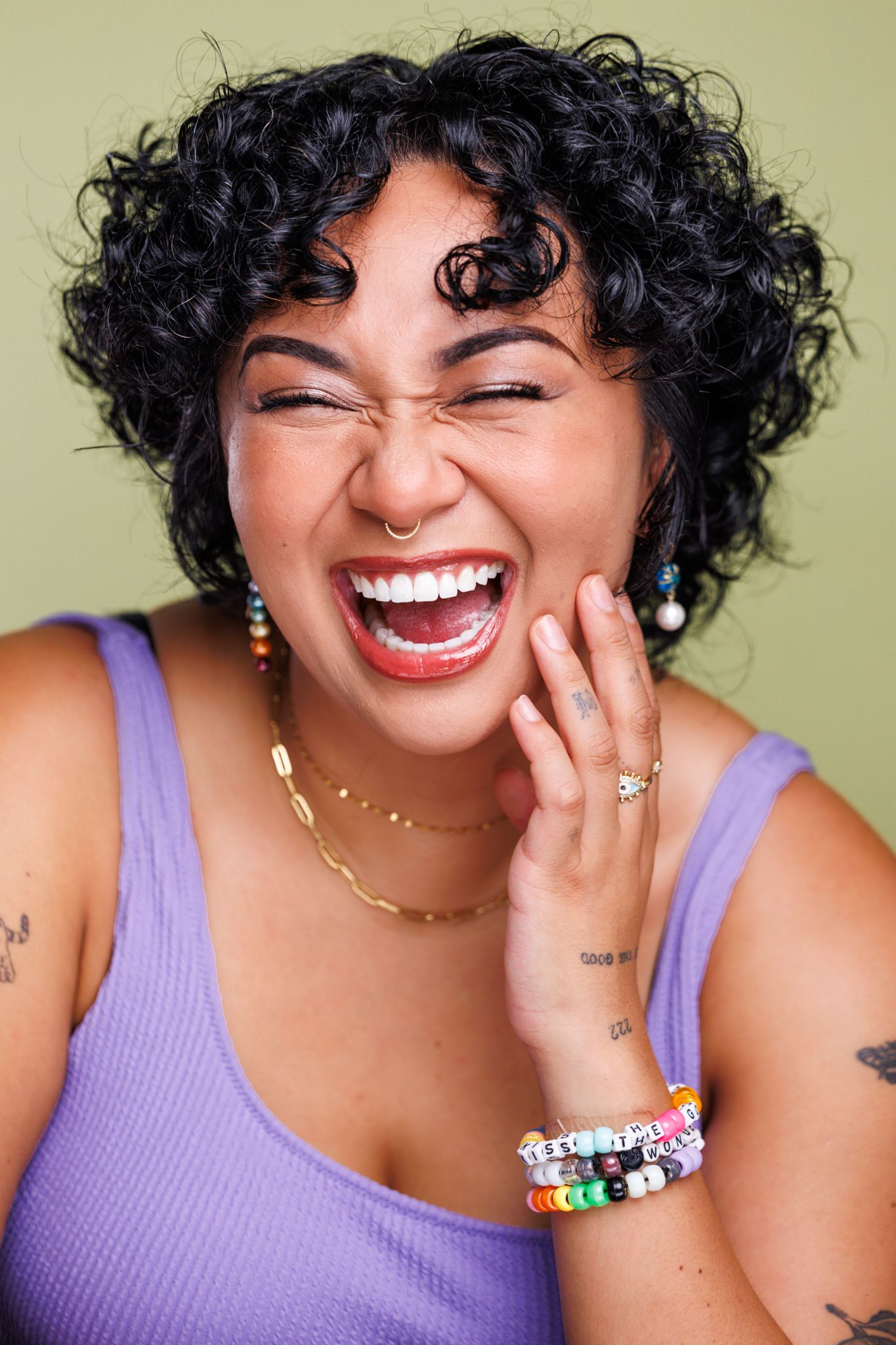 Portrait of a woman with curly hair wearing purple, laughing joyfully against a green background.