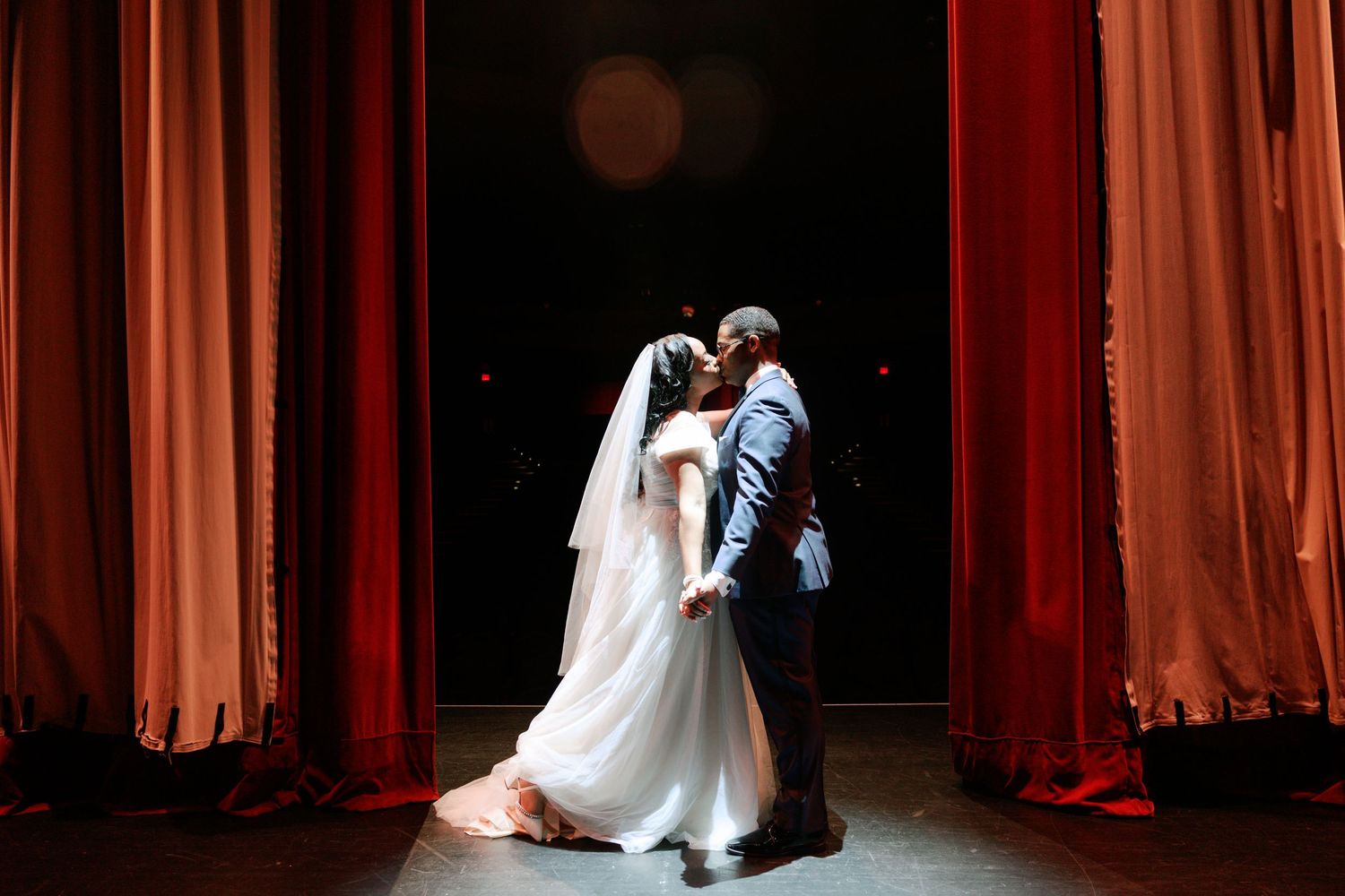 Romantic wedding couple dancing in spotlight with flowing red curtains behind them.
