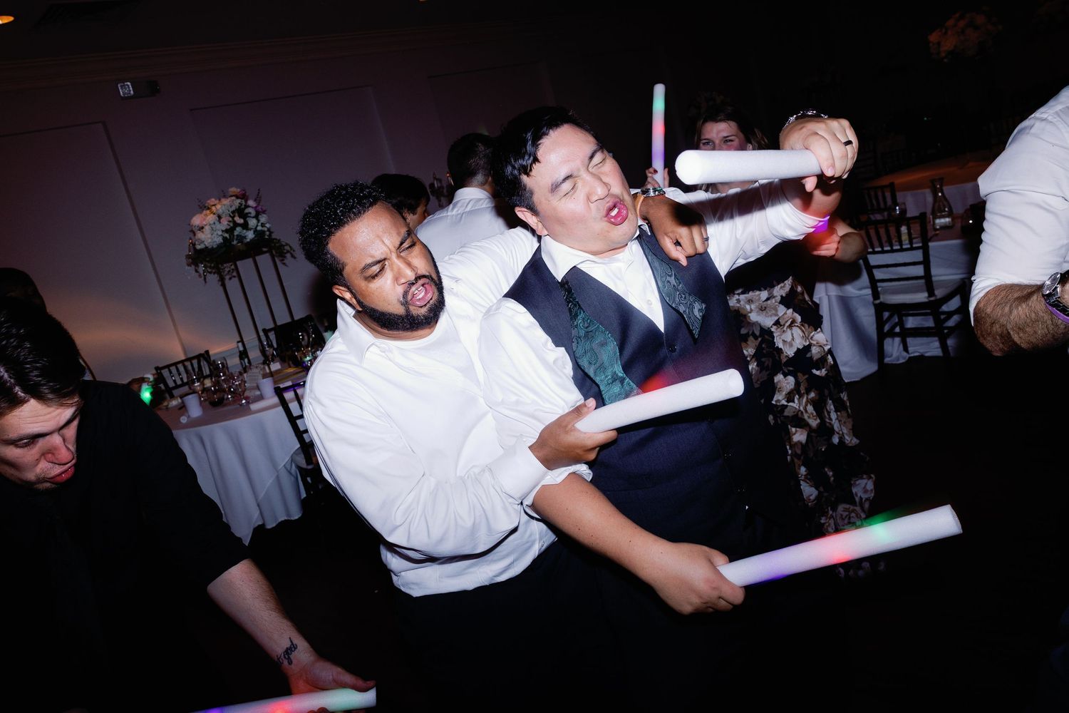 Wedding party guests wave colorful LED foam sticks while dancing in a dark reception venue.