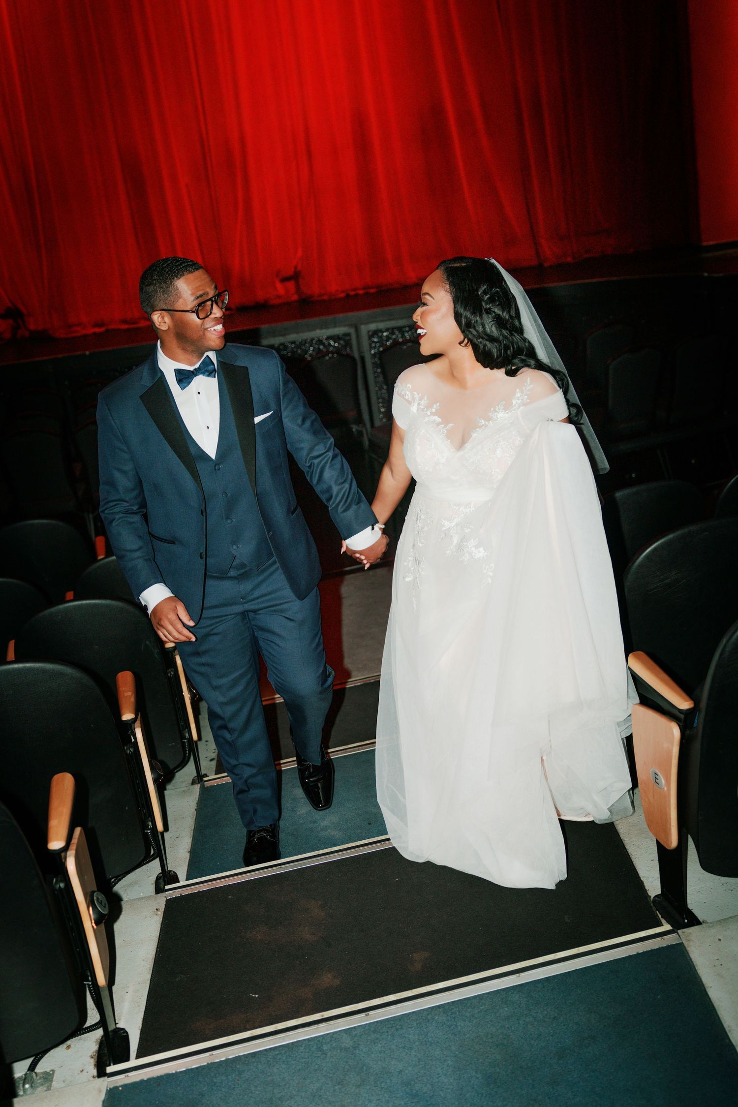 Couple in wedding attire walking through theater seating area with red curtain backdrop.