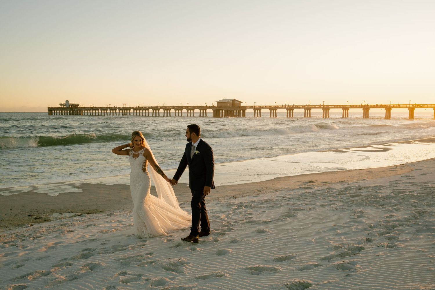 Wedding couple strolls along beach with pier silhouetted against sunset in background.