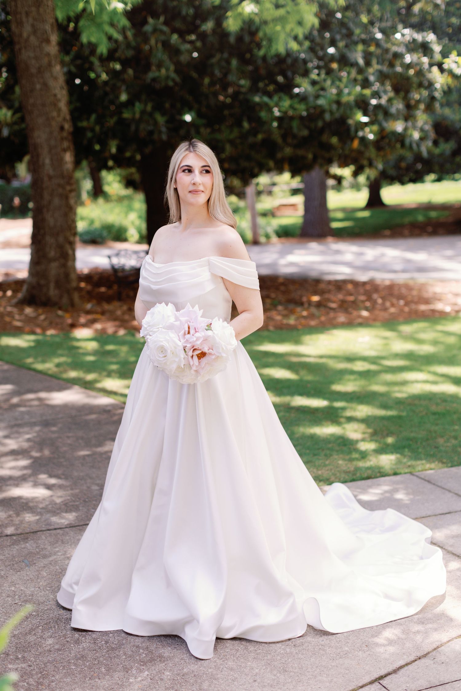 A beautiful white wedding gown flows elegantly across a garden pathway in natural sunlight.