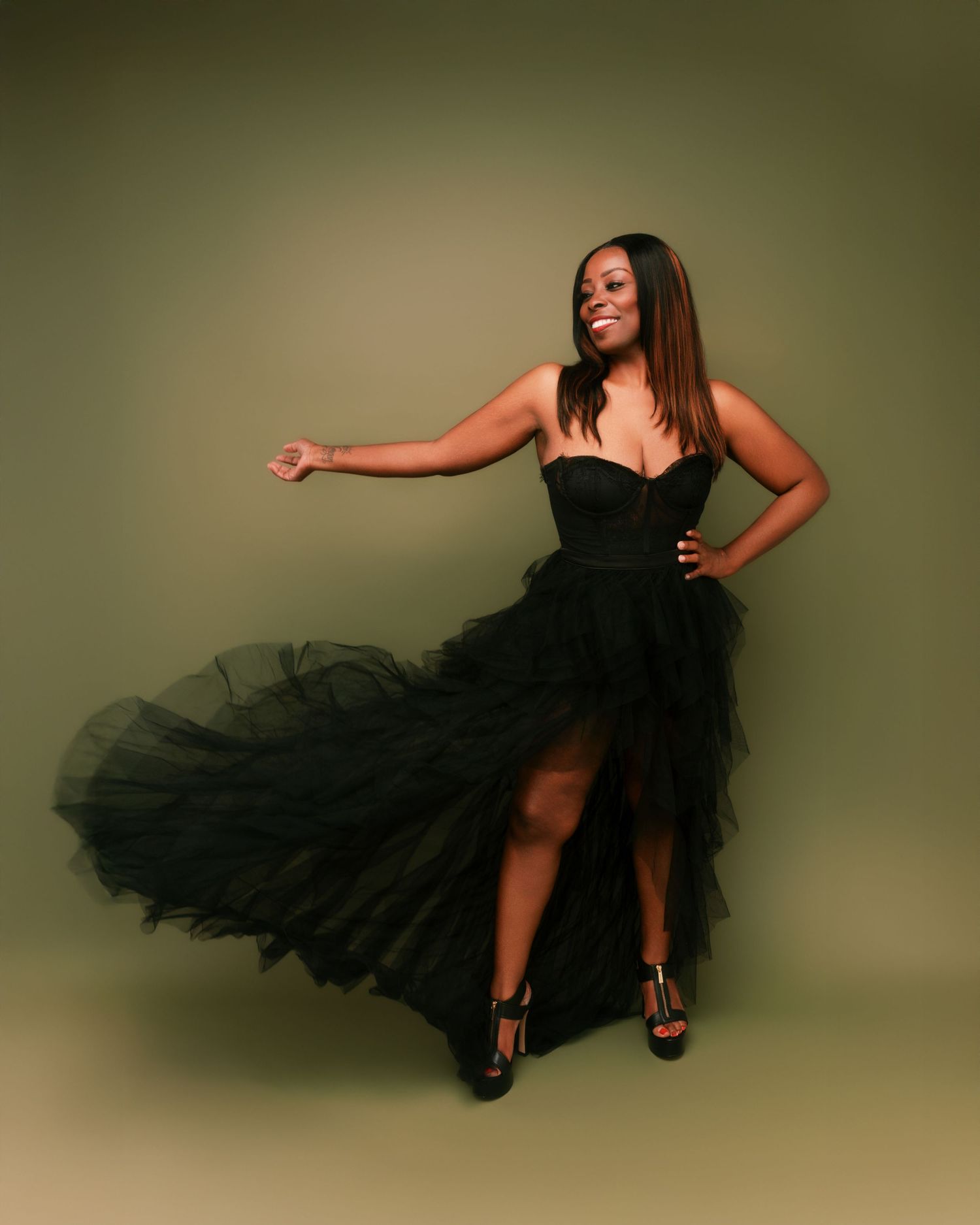 A model poses in a flowing black tulle dress with one leg extended against an olive background.