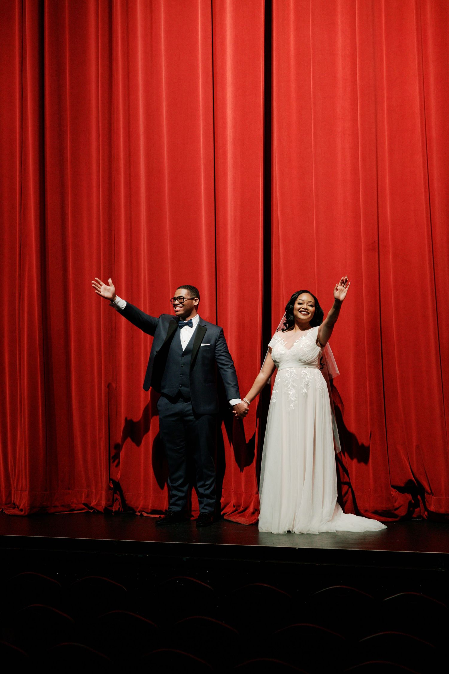 Wedding couple poses triumphantly against dramatic red theater curtains on stage.