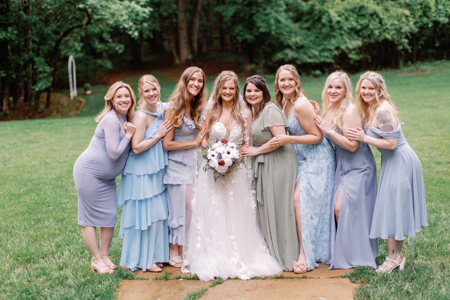 Bridesmaids in soft blue dresses pose together on a grassy lawn during a summer wedding celebration.