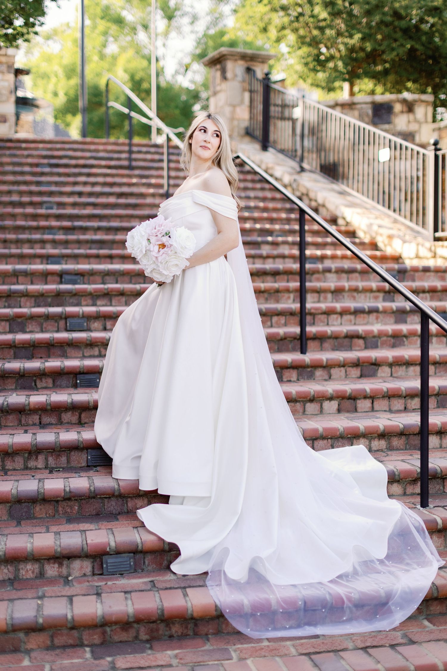 Bride in flowing off-shoulder gown twirls on brick staircase while holding white bouquet.