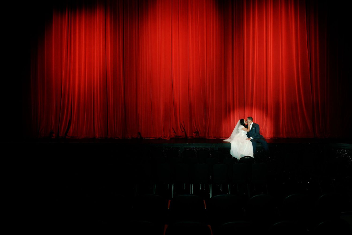 Dramatic wide shot of wedding couple on dark theater stage with red curtain.